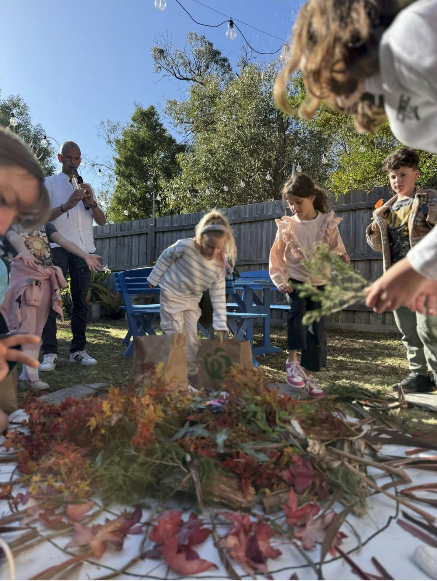 Children and an adult gather outdoors under a clear blue sky, with trees and string lights in the background, participating in an autumn-themed activity involving colorful fall leaves and natural objects on a table.