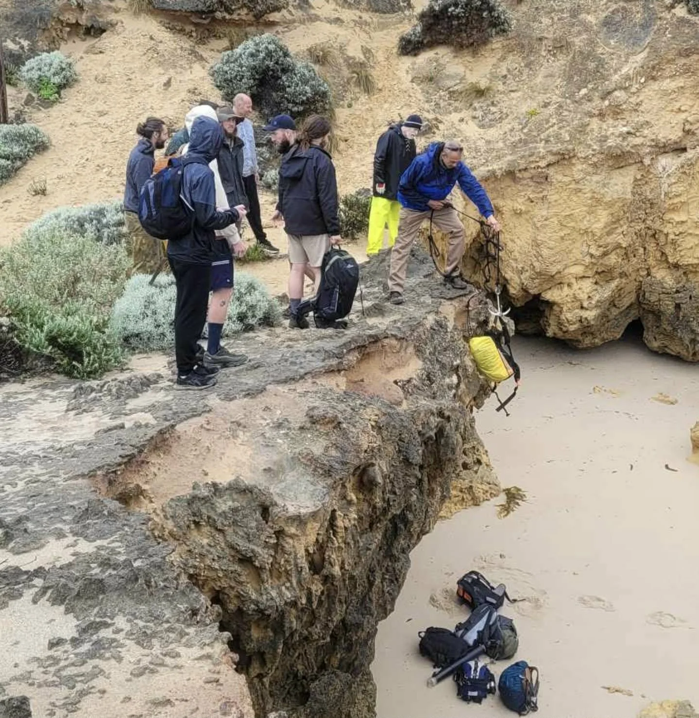 Group of people on rocky ledge overlooking sandy beach, some appear to be preparing for a canyon or cliff descent with gear.