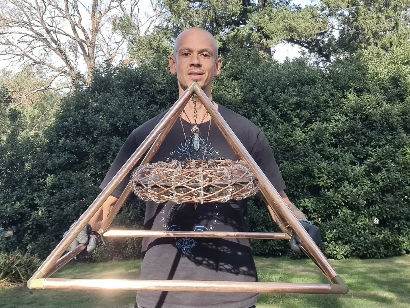 A person standing outdoors holding a large, triangular metal frame with a woven decorative piece suspended in the center. The background features trees and greenery.