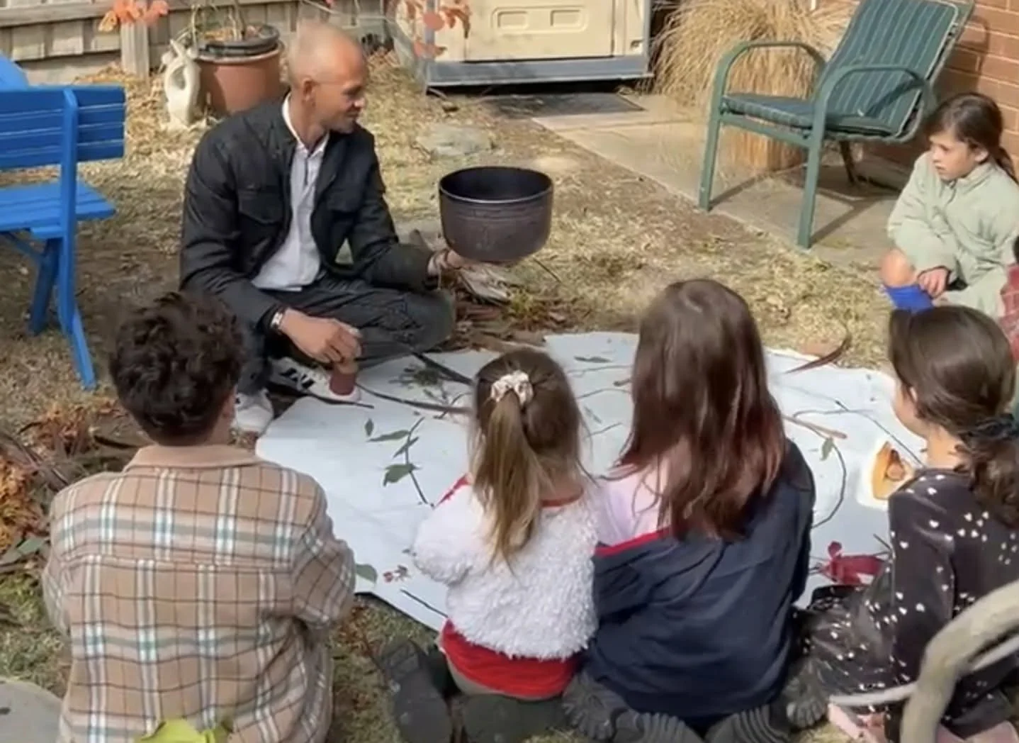 A man appears to be teaching children about nature outdoors, showing a small plant or twig. The children are seated on the ground around him on a white cloth, with some wearing jackets and sweaters, indicating cooler weather. There are outdoor chairs