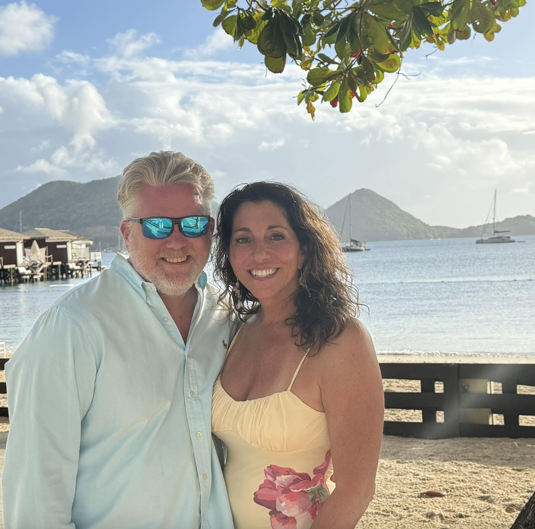 A couple posing for a photo on a beach with a mountain, boat, and wooden overwater bungalows in the background. The man is wearing a light blue shirt and sunglasses, and the woman is in a yellow dress with pink floral patterns. There is a tree branch overhead with green leaves.