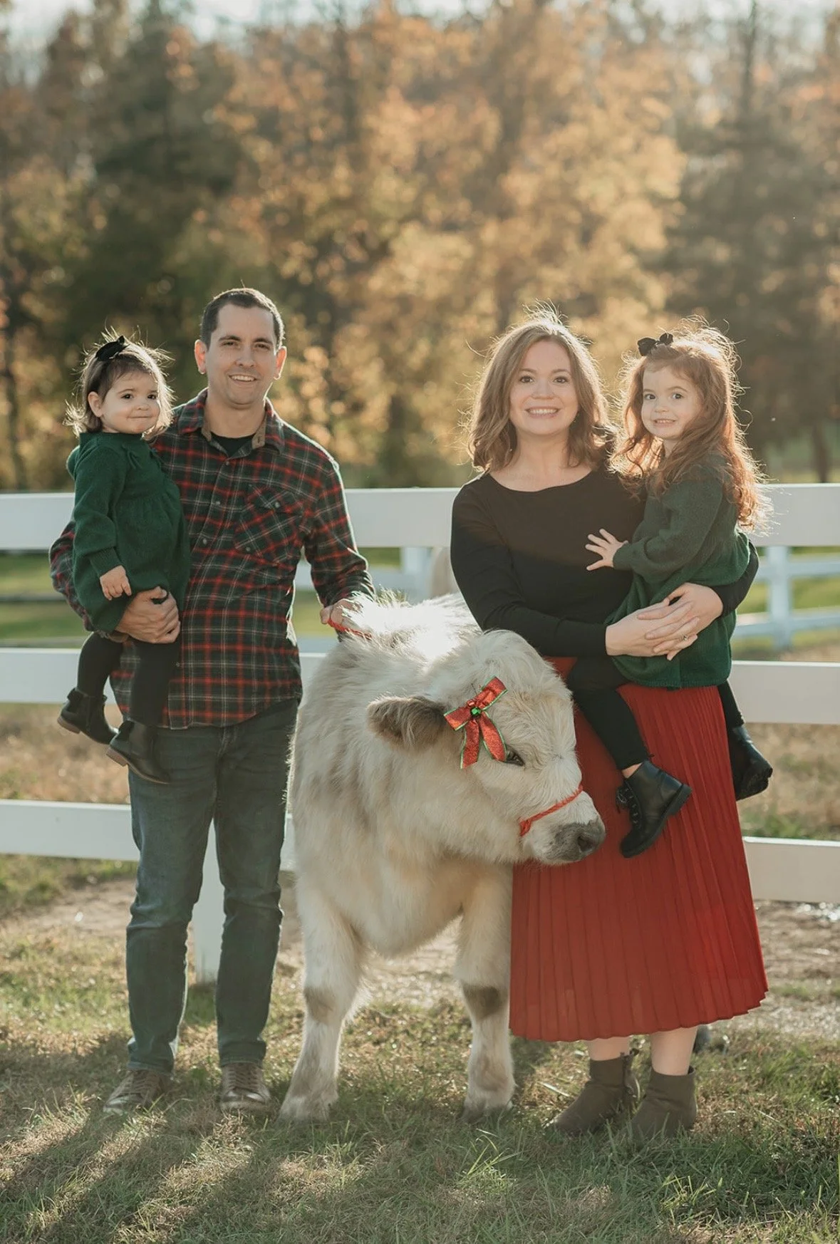A family of four posing outdoors with a large white dog decorated with a red bow and collar. The father and daughter are on the left, with the daughter being held by the father. The mother and younger daughter are on the right, with the daughter sitting on the mother's lap. The background features trees with autumn foliage and a white fence.