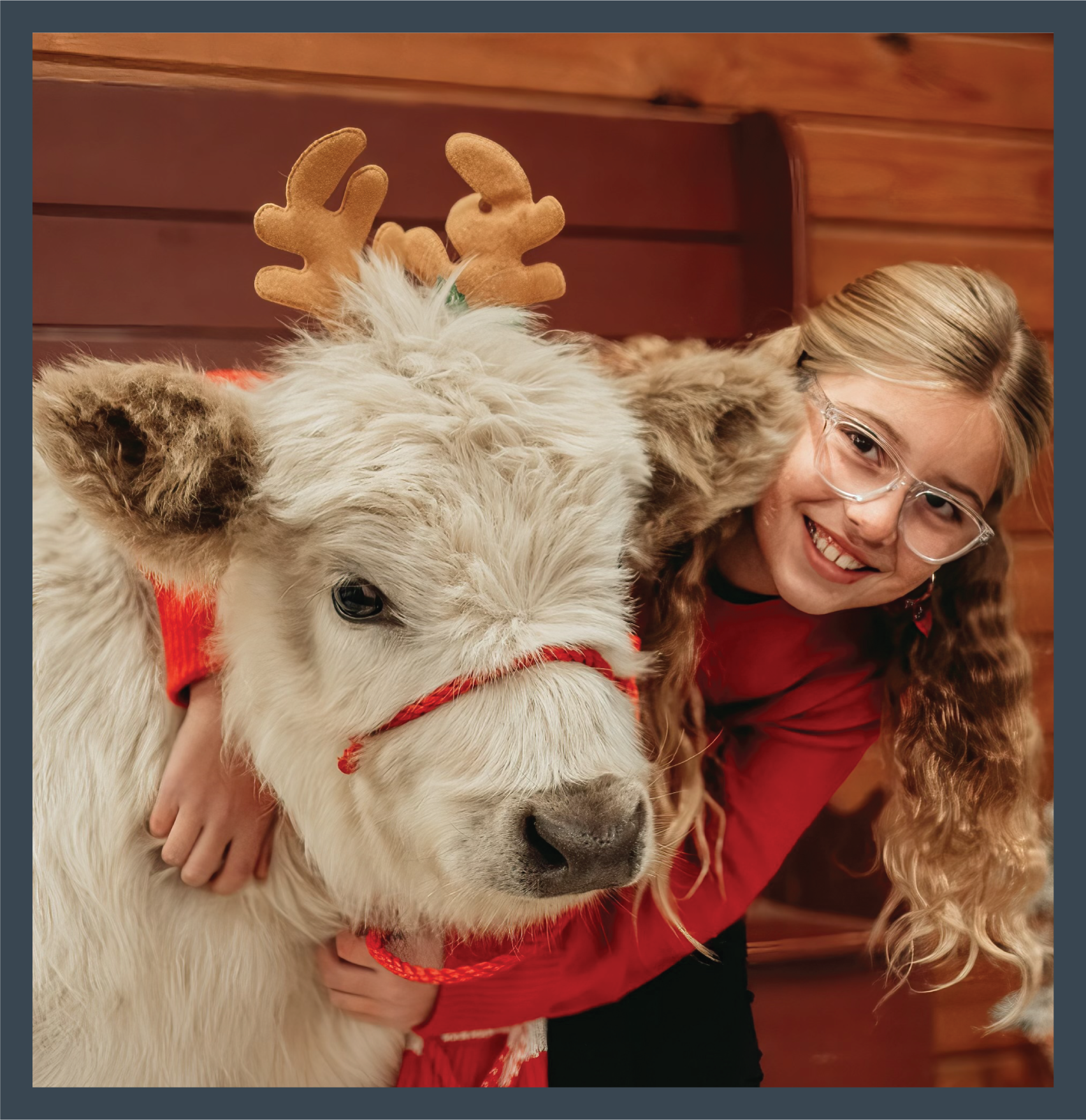 A joyful woman with glasses hugging a white dog wearing reindeer antlers, both indoors with a wooden wall background.