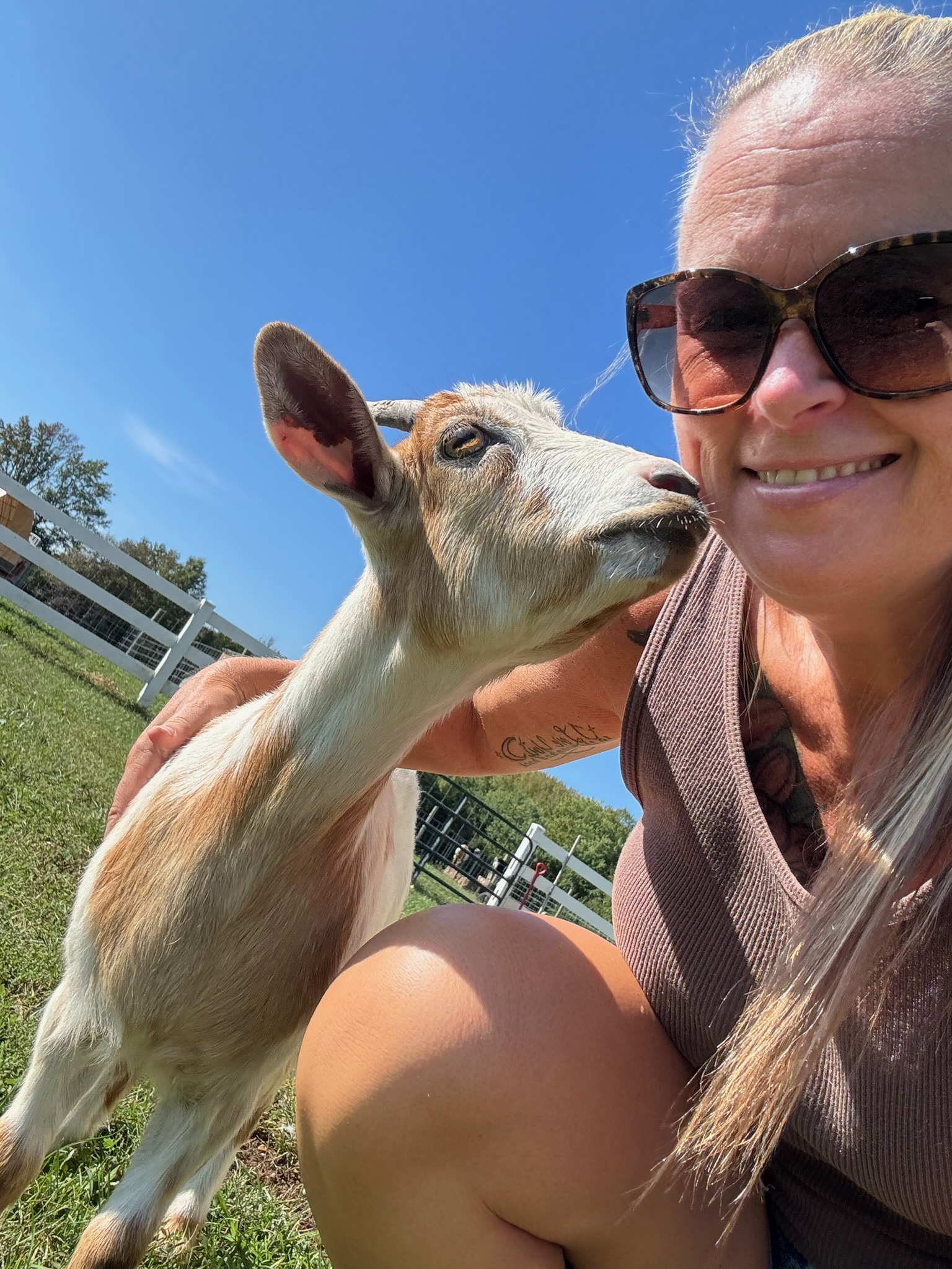 A woman wearing sunglasses smiling next to a calf in a grassy outdoor area under a blue sky.