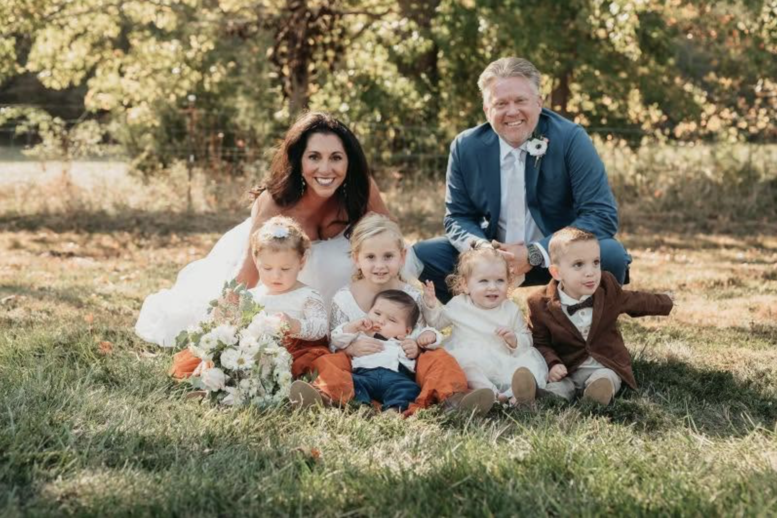 A family of eight posing outdoors on a grassy field during autumn. The group includes a woman in a wedding dress, a man in a blue suit, and six young children dressed in formal attire. The background features trees with fall foliage.