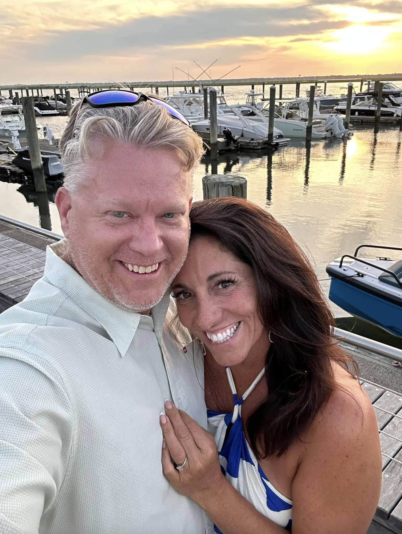 A smiling man and woman taking a selfie on a dock near boats at sunset.