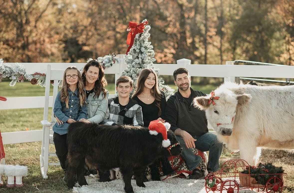 Family of five in Christmas outfits sitting on a bench outdoors with a small Christmas tree, festive decorations, and two calves wearing Christmas hats.