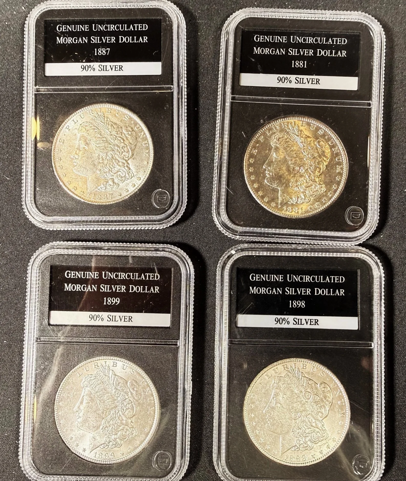 Four Morgan silver dollar coins in plastic holders with labels indicating they are genuine, uncirculated, 90% silver, from years 1887, 1881, 1899, and 1898.