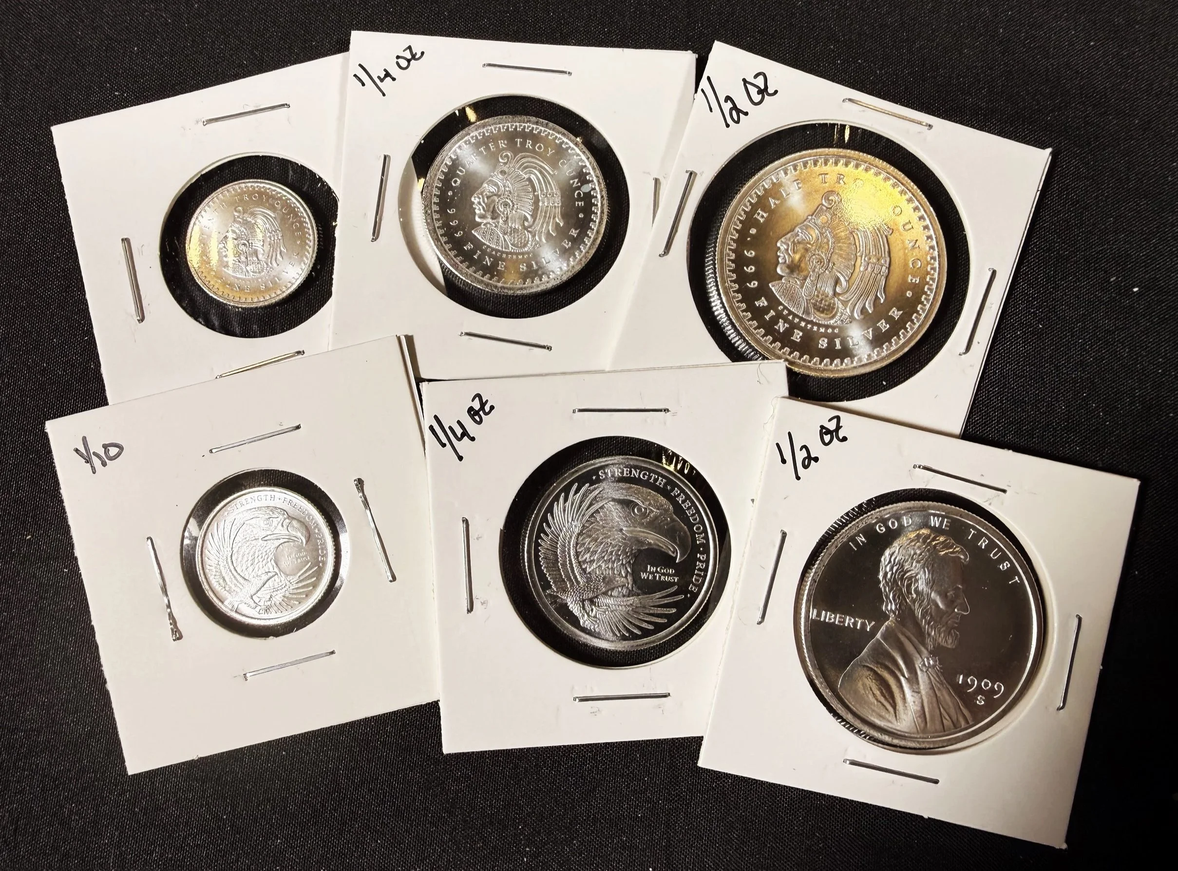Three silver coins in protective holders with white paper inserts and handwritten labels indicating their weights, Set 1/4 ounce, 1/2 ounce, and 1 ounce.