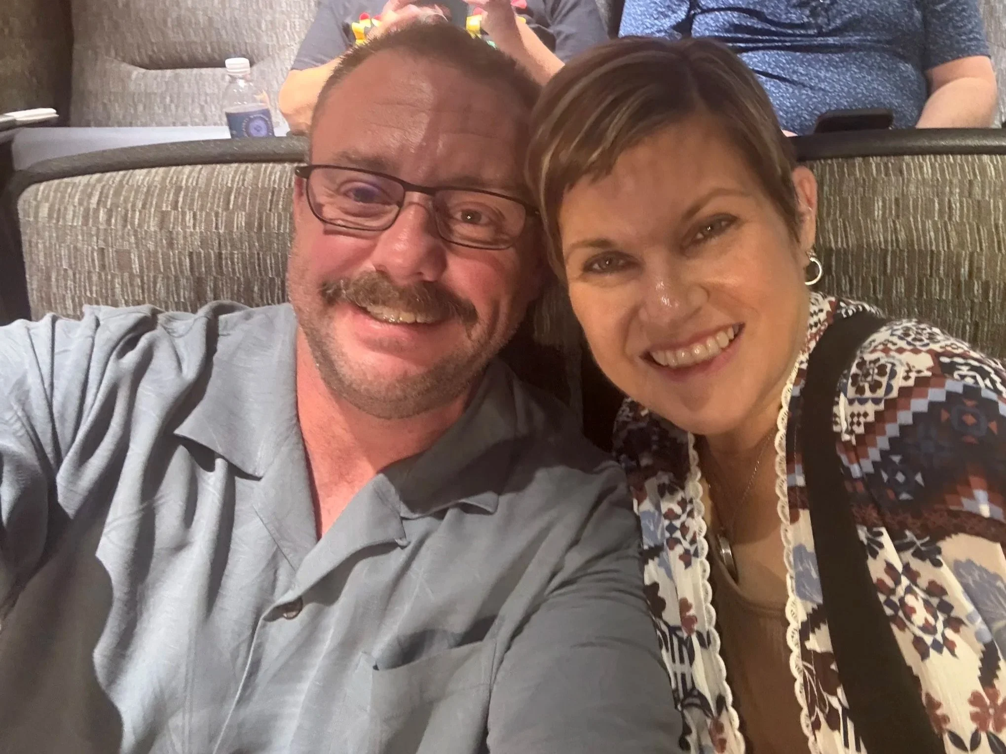 A man with glasses and a woman with short brown hair smiling as they take a selfie together in a row of theater seats. The background shows other people seated behind them, some with drinks and snacks.