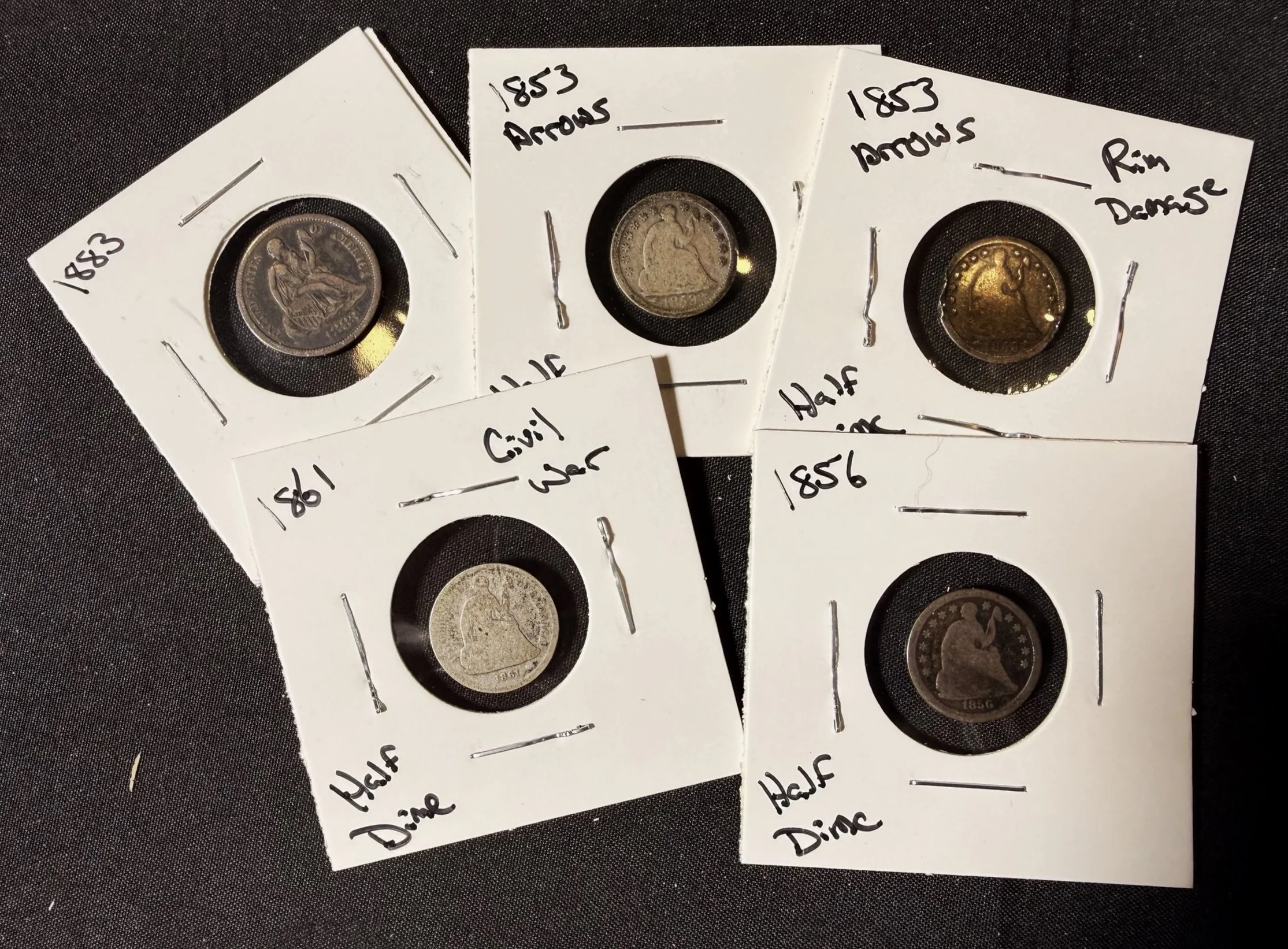 Three old coins in white cardboard coin holders with handwritten labels. The labels include dates and descriptions such as '1853 Arrows,' 'Half,' and 'Dime.' The coins are displayed on a black textured surface.