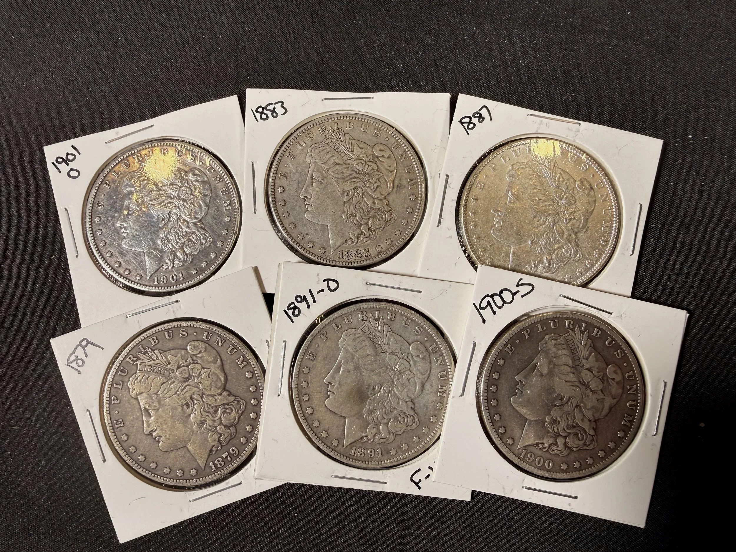 A collection of six old silver coins, each labeled with the year of minting, including 1901, 1883, 1887, and 1900, placed on a dark surface and protected in individual white coin holders.