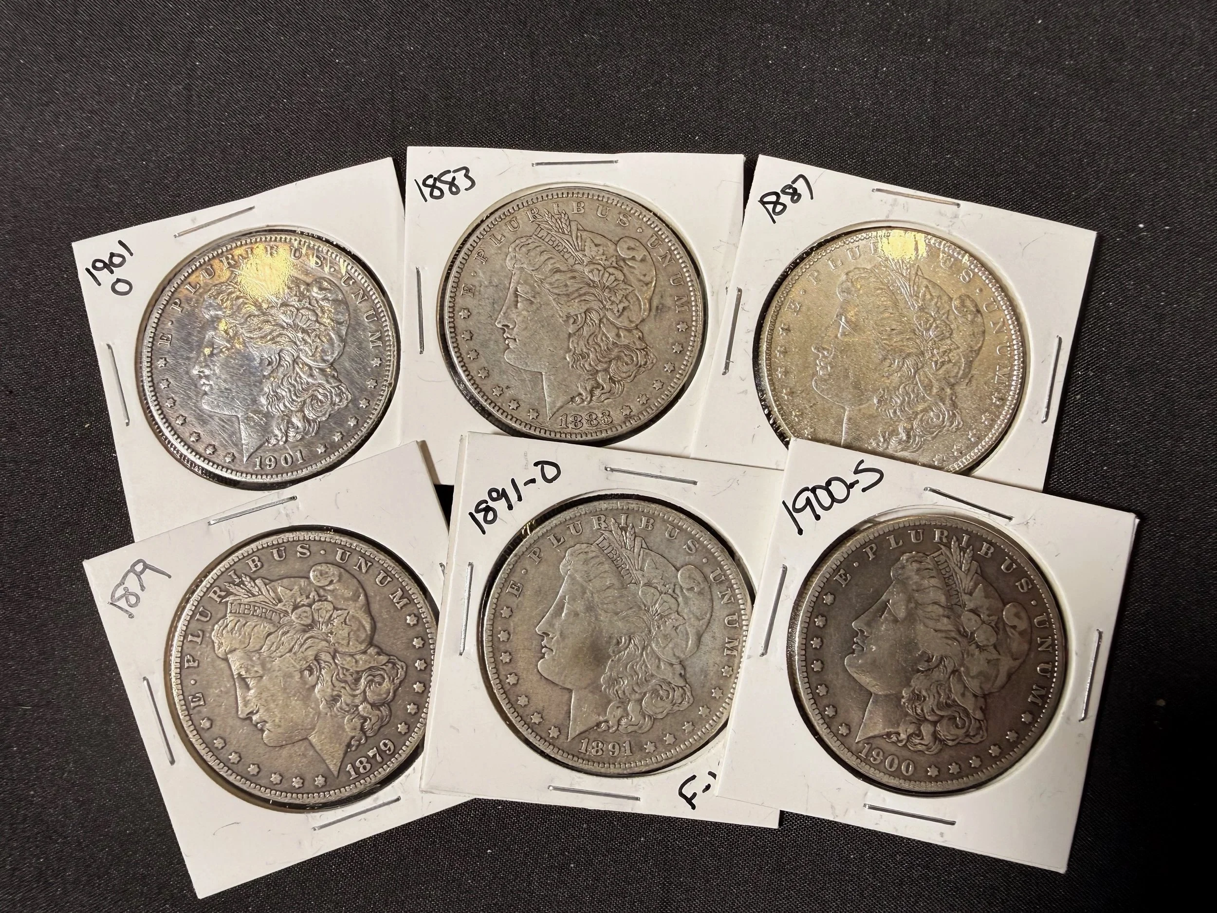 A collection of five silver coins in plastic holder with handwritten year labels: 1901, 1883, 1887, 1891, and 1900, displayed on black fabric background.