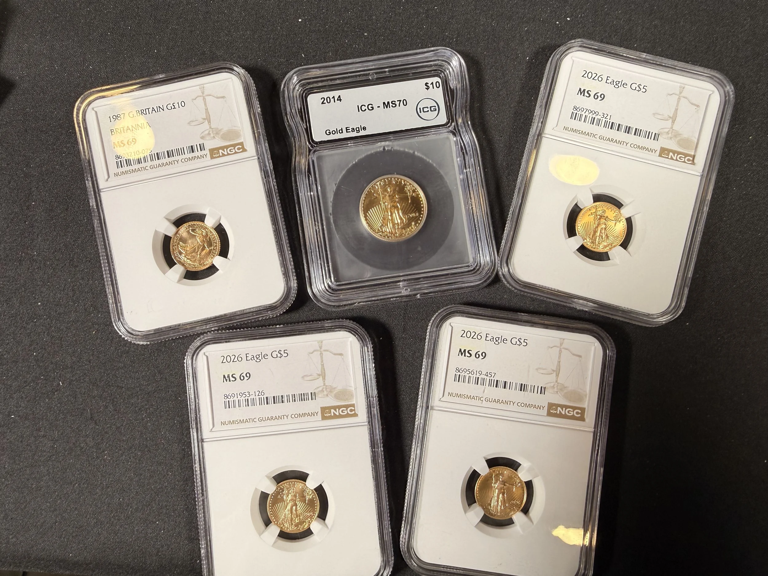 Close-up photo of three gold coins in protective cases, each labeled with year, type, and grade, placed on a black surface.