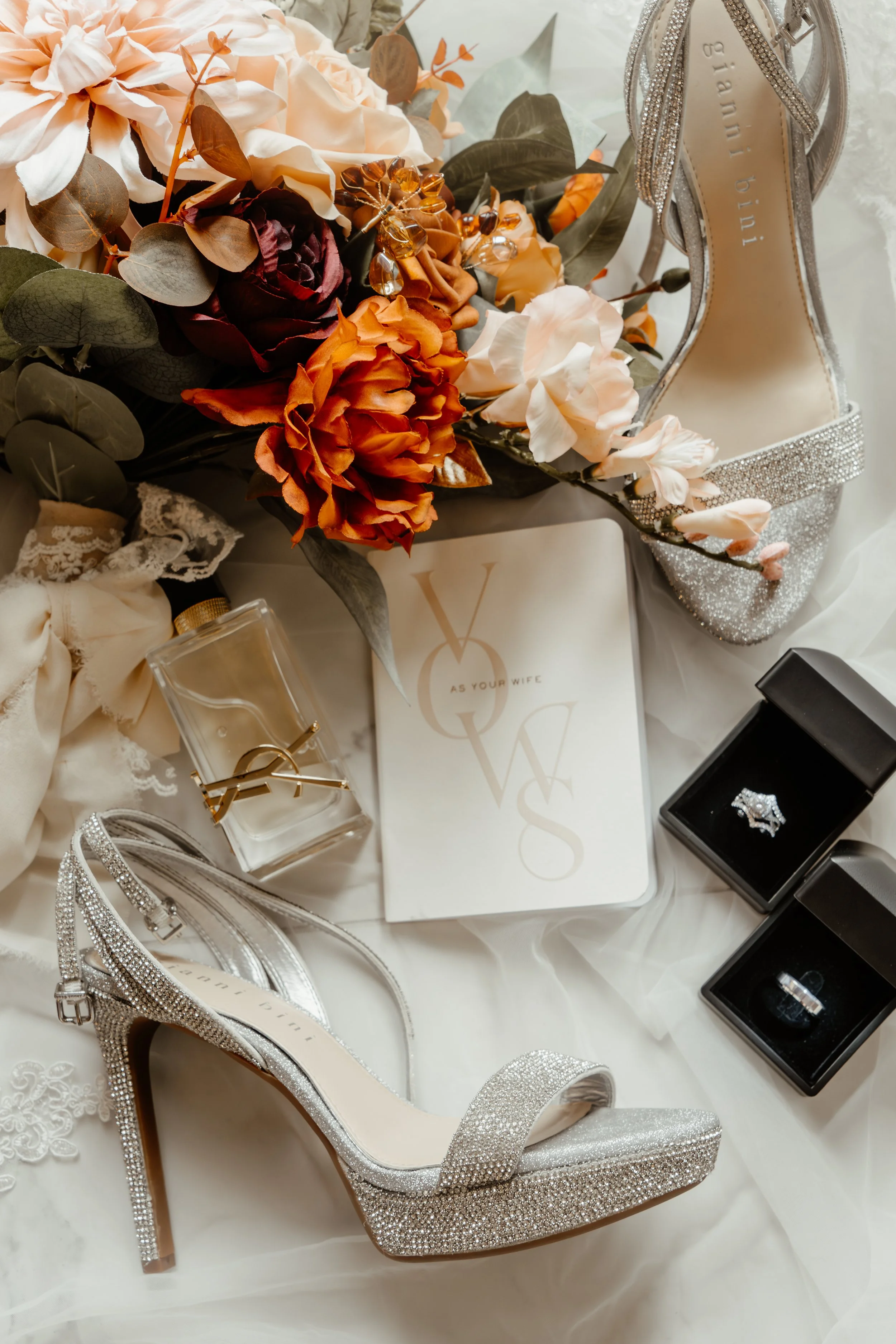 Silver, glittery high-heeled shoes surrounded by a bouquet of peach, coral, and burgundy flowers, along with jewelry items, perfume bottle, and gift boxes, likely for a wedding or special occasion.