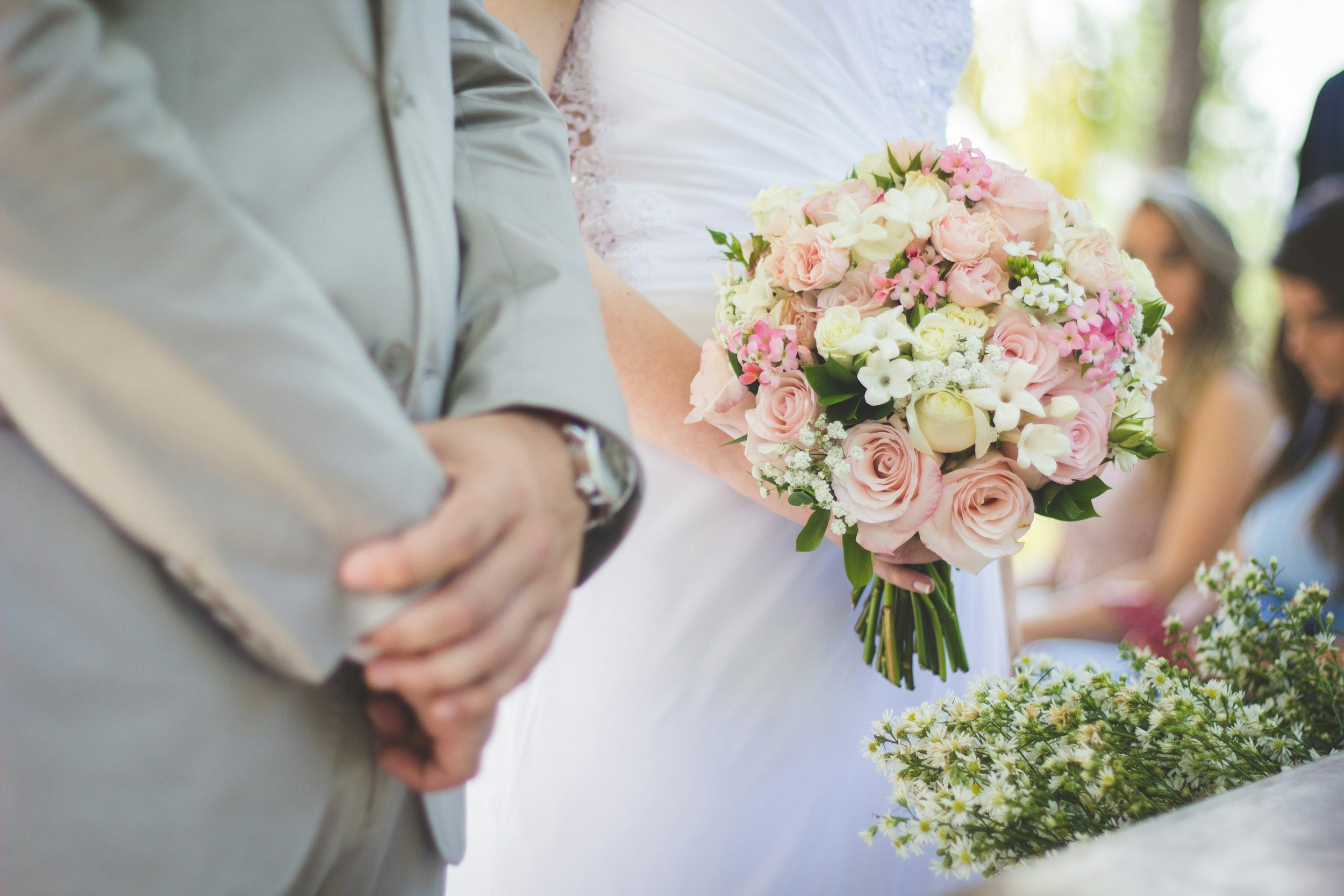 Close-up of a wedding bouquet with pink and white roses and small white flowers, held by a bride. The groom is standing nearby, wearing a gray suit, with guests blurred in the background.