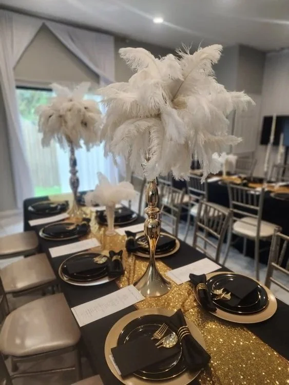Elegant table setting with black and gold plates, black napkins, gold chargers, decorated with tall centerpieces of white feathers on gold stands, set for a formal event or celebration.