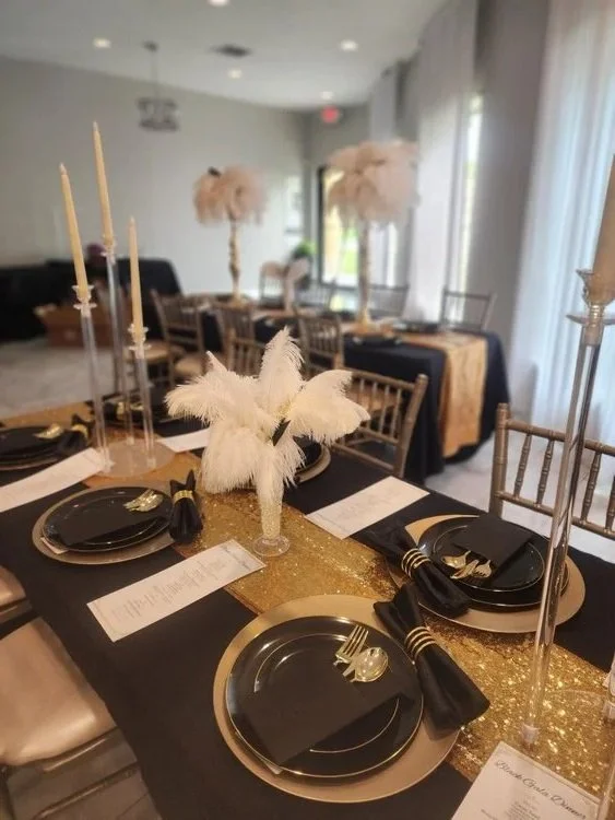 Elegant dining table setup with black and gold plates, black napkins, gold cutlery, tall gold candlesticks, and white feather centerpieces at a formal event or wedding reception.