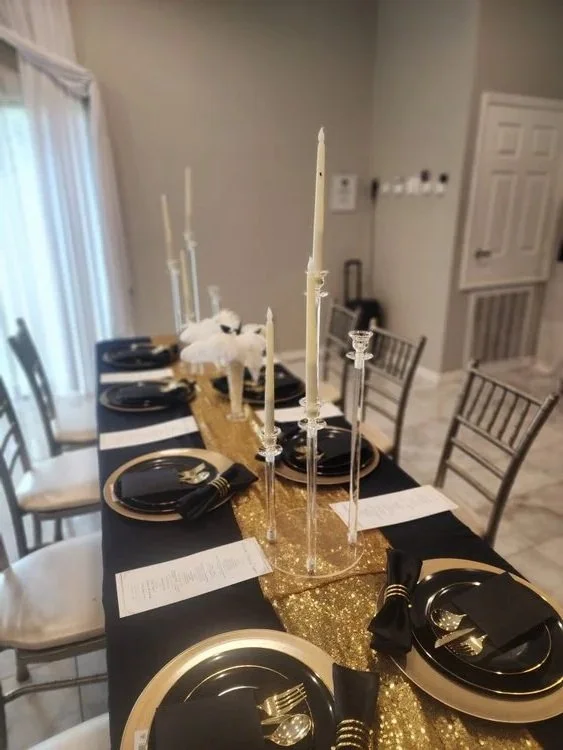 A decorated dining table with black and gold plates, black napkins with gold rings, gold chargers, and a gold glitter table runner. Tall white candles in clear candle holders are as a centerpiece, with some candles unlit. The table is set for a celeb