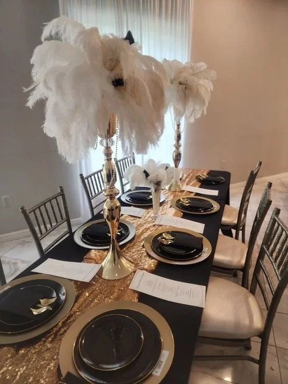 A dining table set for a formal event with black plates, gold chargers, and black napkins. The table has a gold sequin table runner and elegant tall gold candle holders with large white feather arrangements on top.