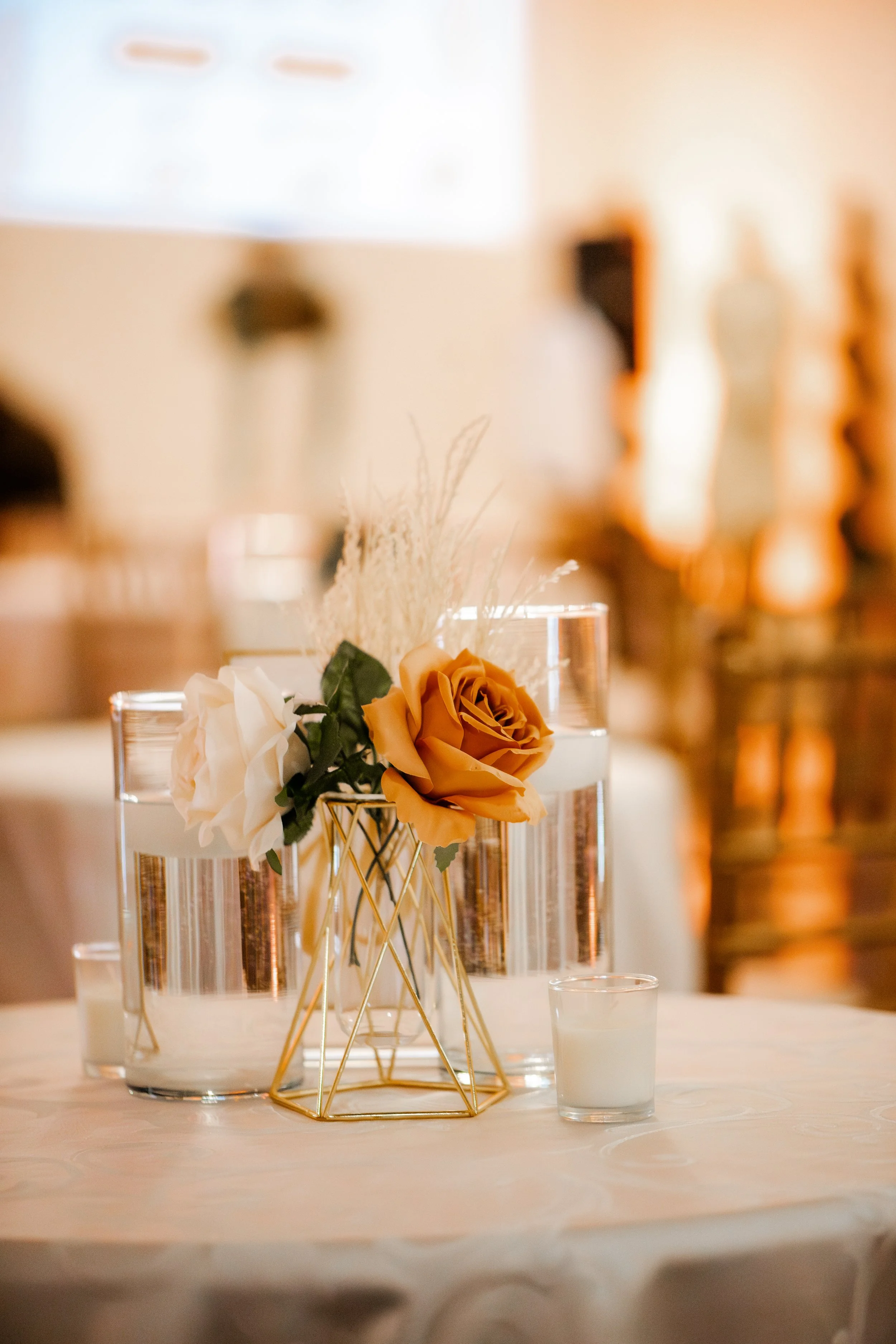Decorative floral centerpiece with white and beige roses, in a gold geometric vase, on a table set for an event or celebration.