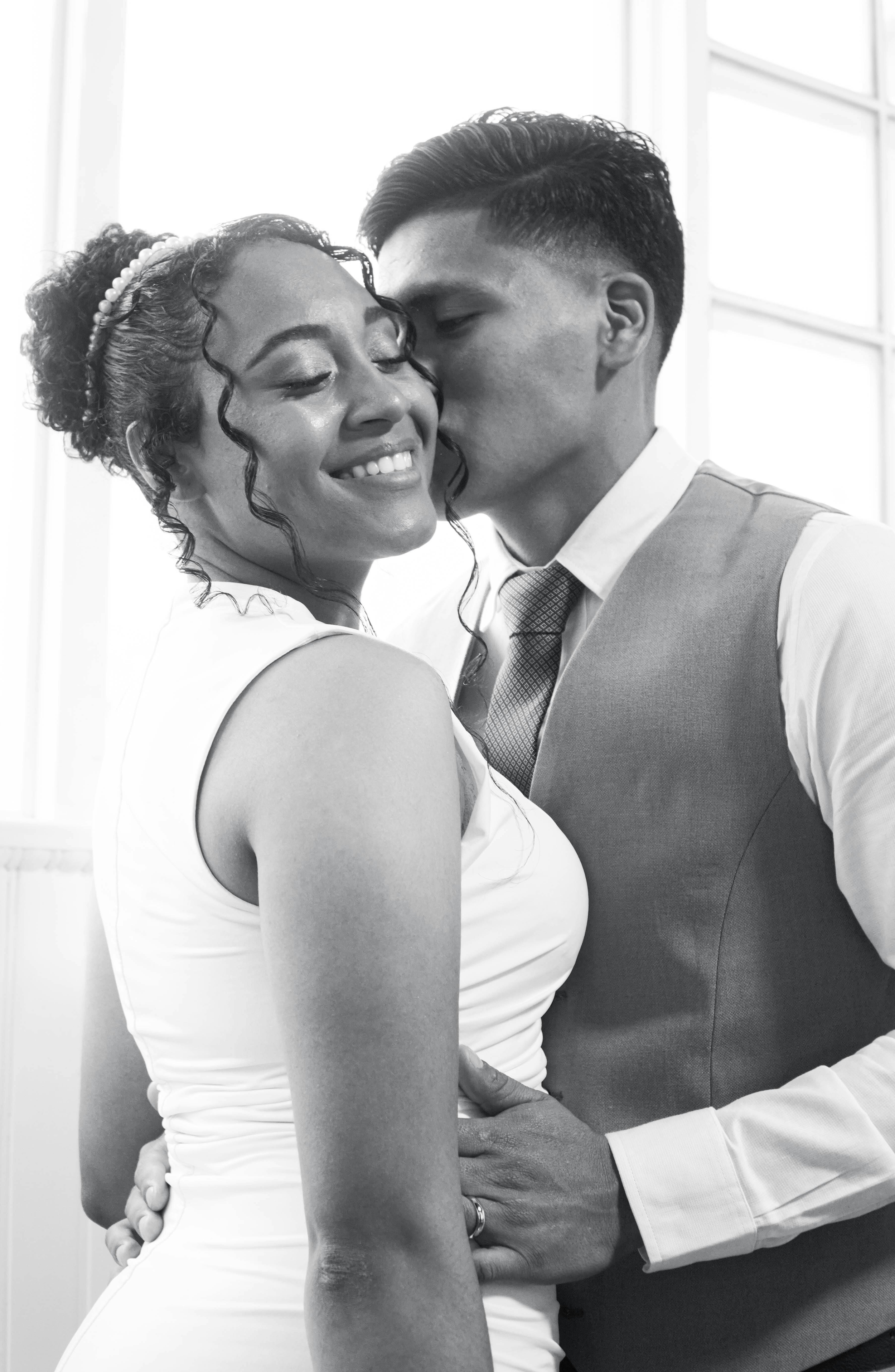 A black and white photograph of a couple with closed eyes, the woman smiling gently as the man kisses her forehead. They are close together indoors near a window.