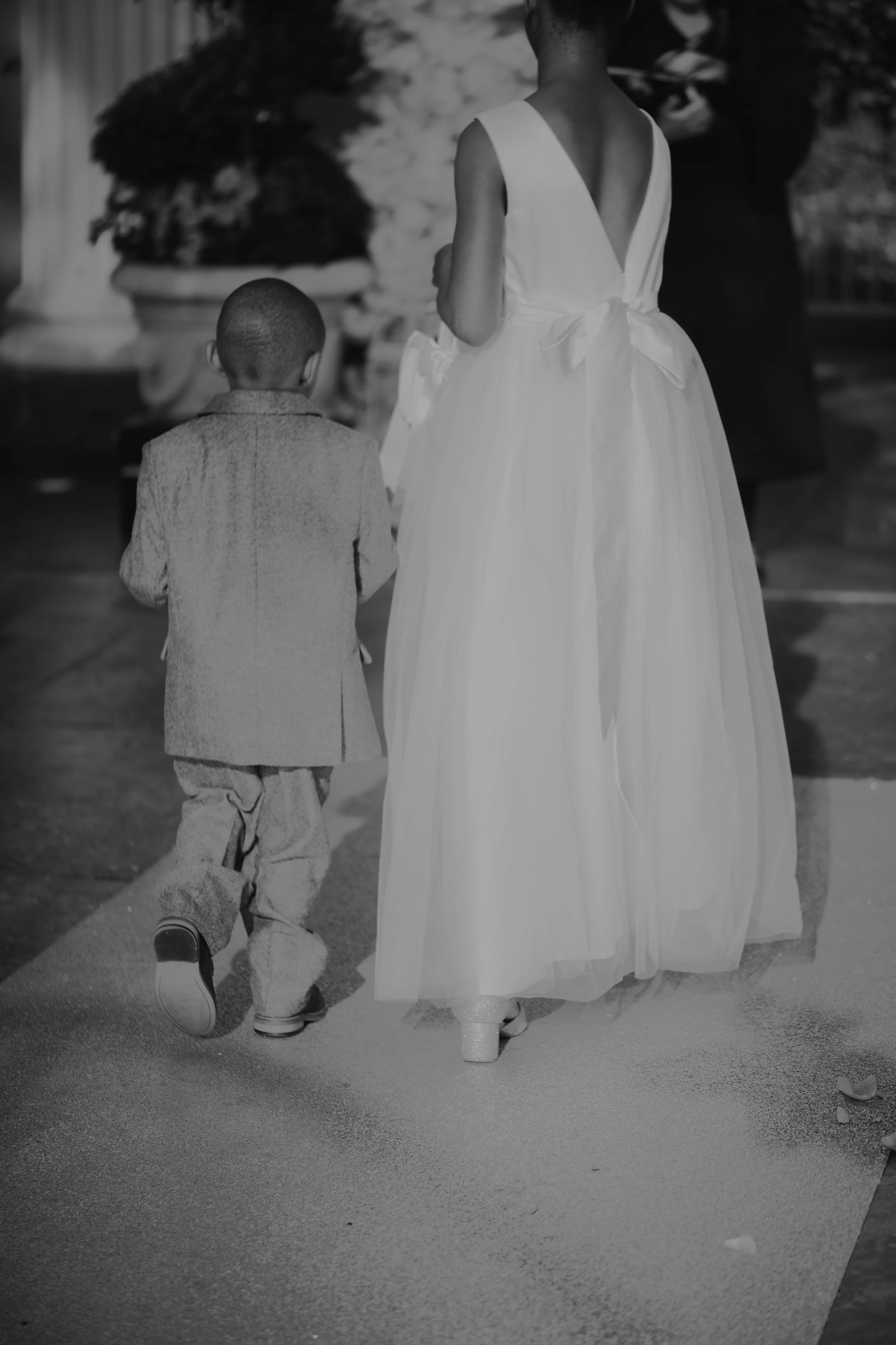A woman and a young boy walking hand in hand on a sidewalk, with the woman in a sleeveless dress and the boy in pants and a jacket.