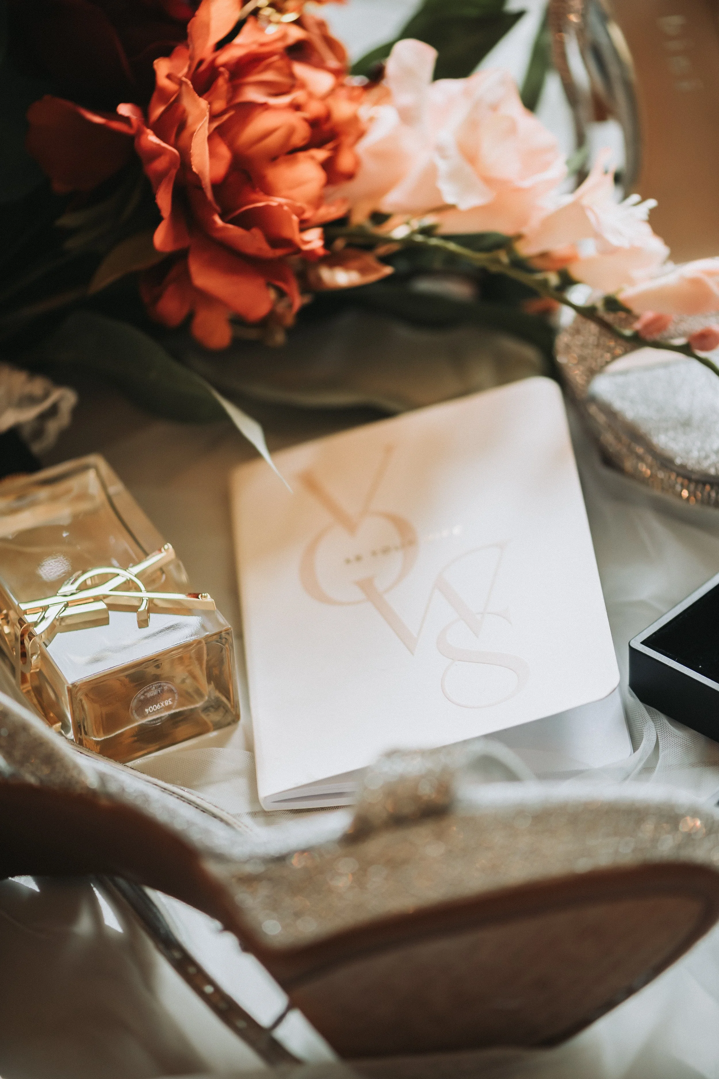 A bouquet of orange and light pink flowers in a glass vase, alongside a white box with gold lettering and a small glass perfume bottle with a gold bow, on a table.