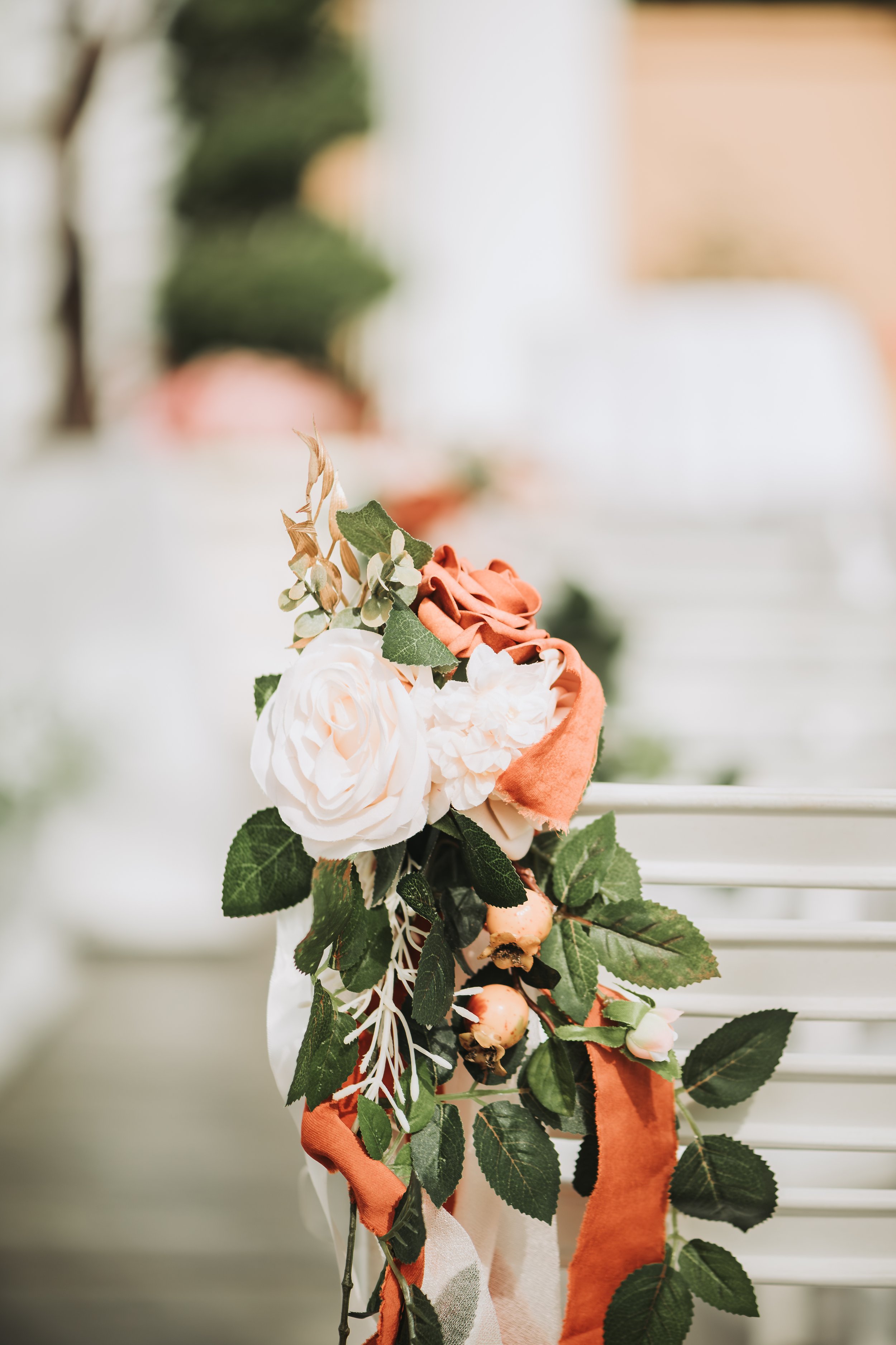 A floral arrangement with white and peach roses, green leaves, and orange ribbons, hanging against a white background.