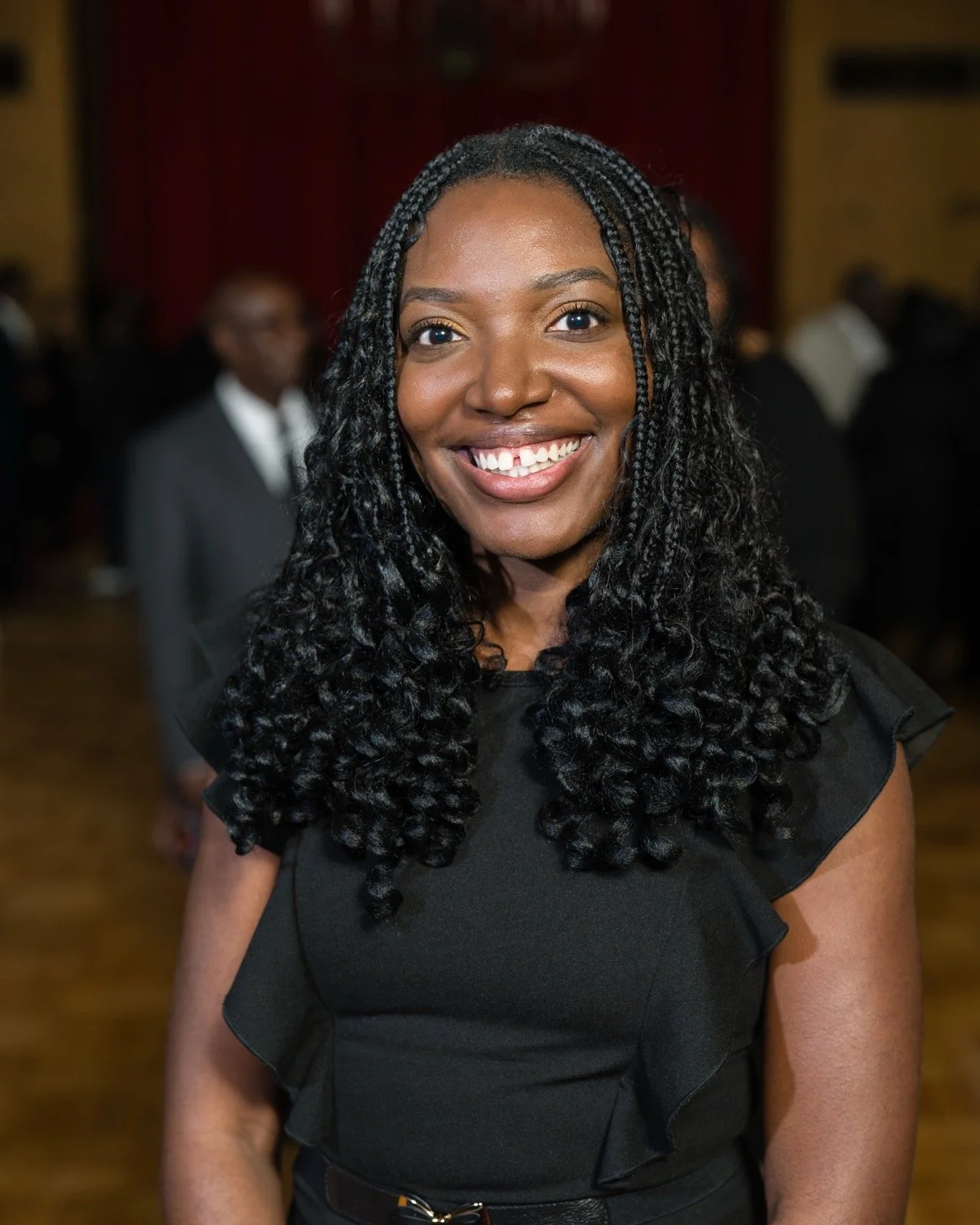 Portrait of a smiling Black woman with braided curly hair wearing a black dress at an indoor event.