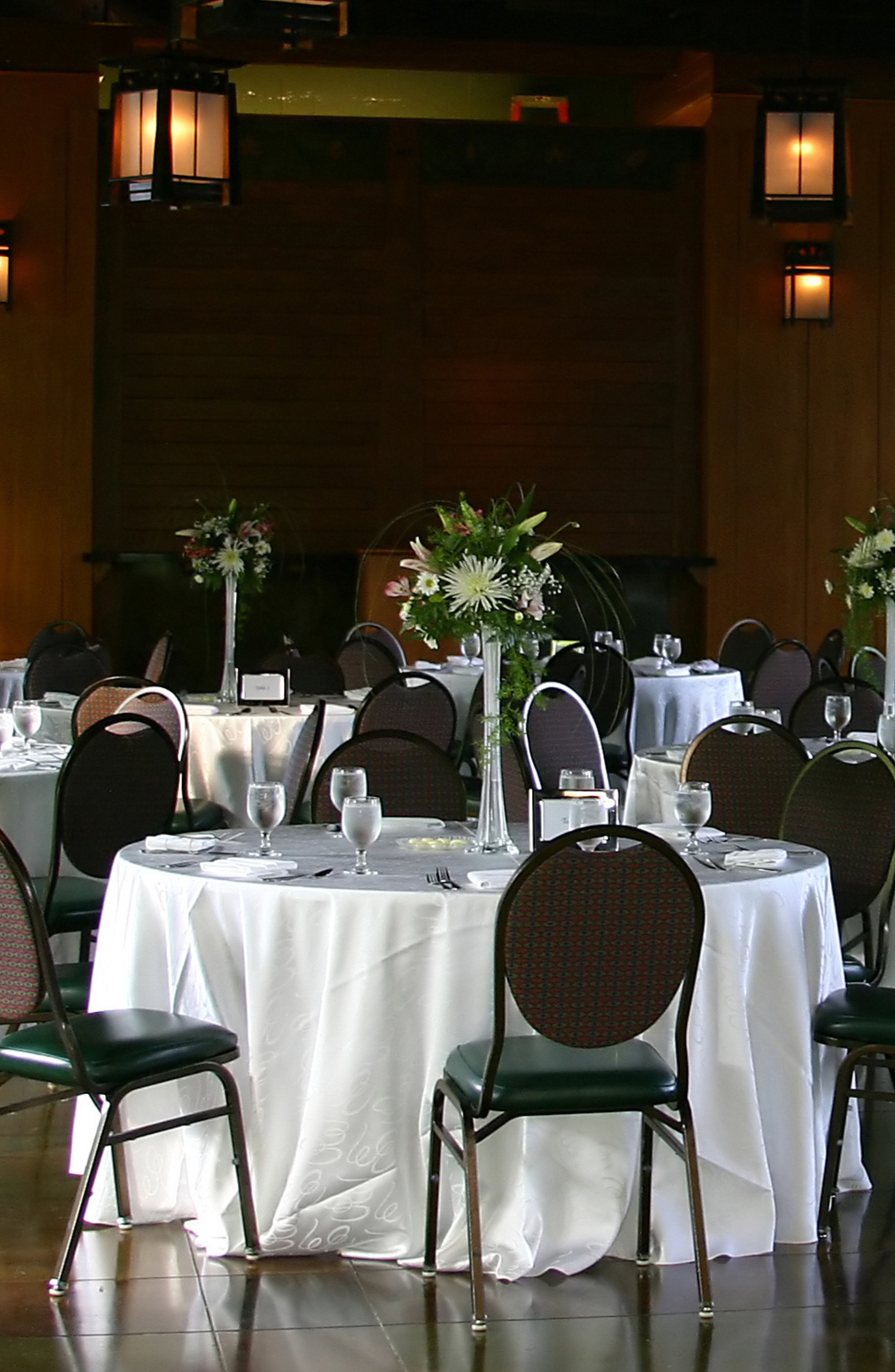 Dining area with round tables covered with white tablecloths, decorated with floral centerpieces, surrounded by black chairs with red patterned backs, set with wine glasses, water glasses, and utensils, in a dimly lit room with dark wood paneling and