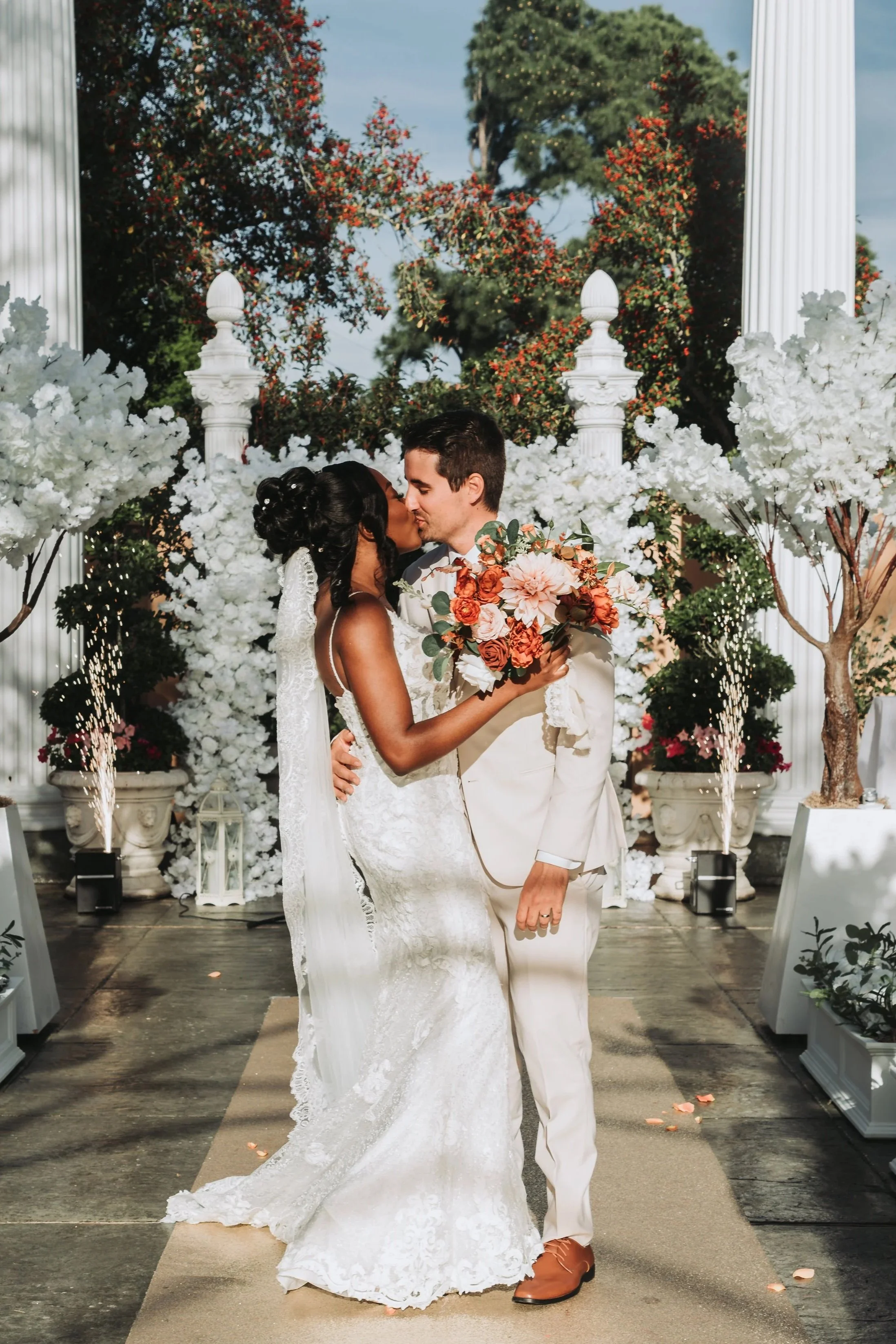 A bride and groom kissing at their wedding ceremony outdoors, surrounded by white floral arrangements and trees, with columns and a decorated aisle.