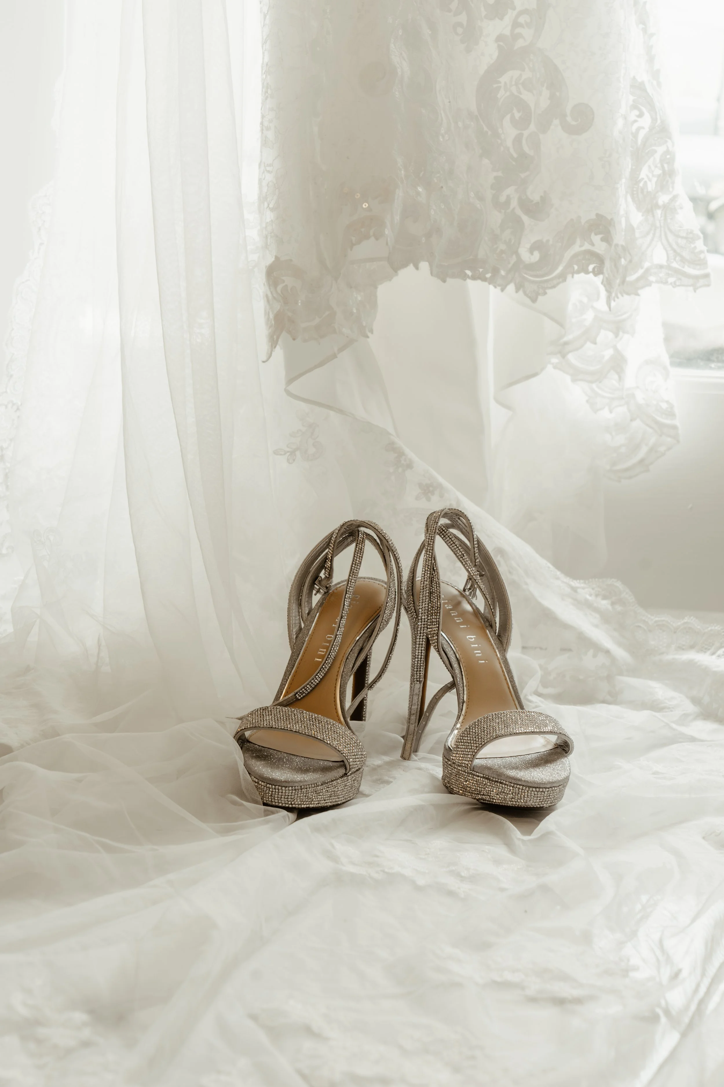 A pair of glittering high-heeled shoes on a white cloth with lace curtains in the background.