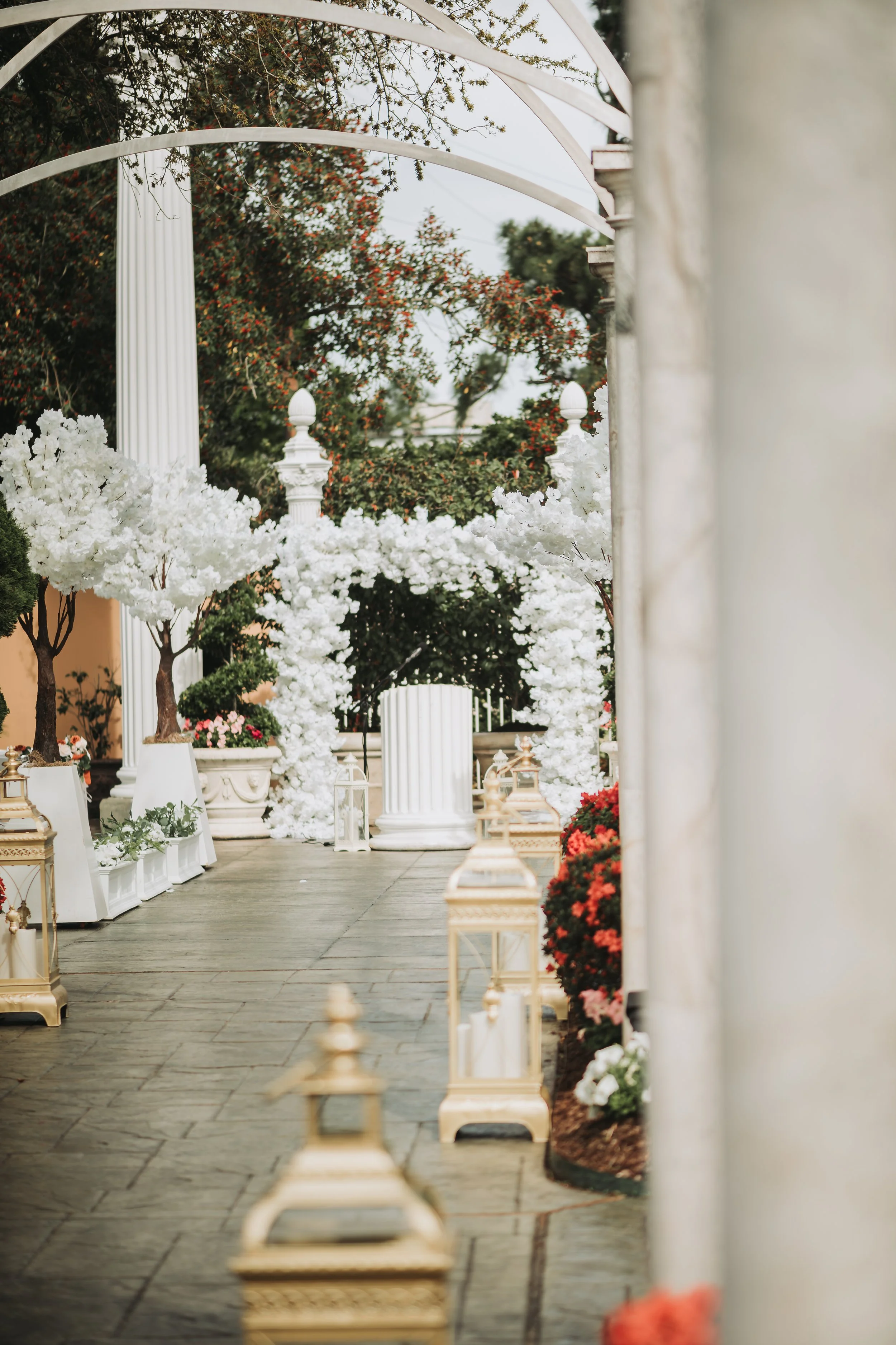 A pathway adorned with white flowers and lanterns, framed by white columns and a decorative arch overhead, in an outdoor garden setting.