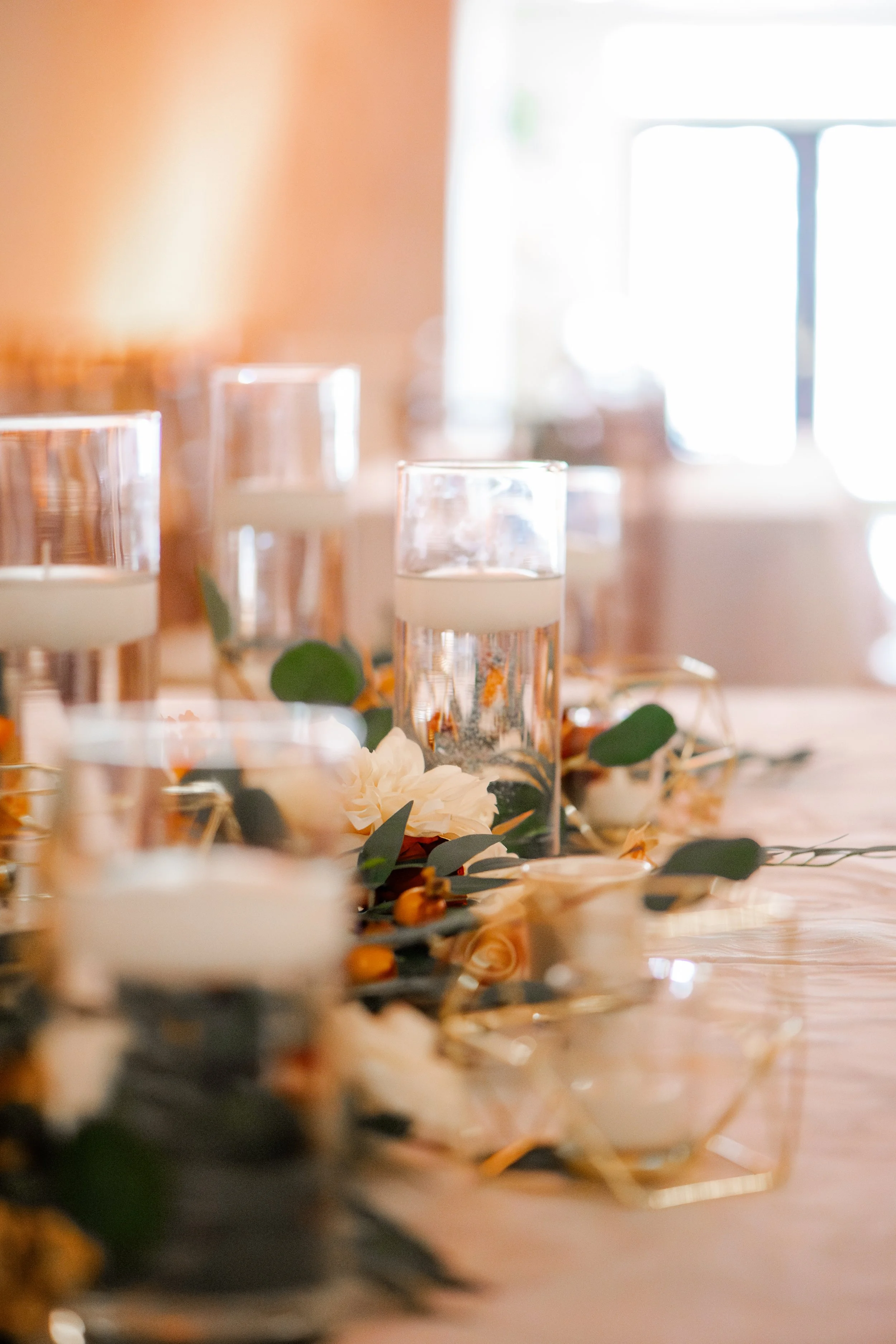 Close-up of a table with tall clear glasses with white candles inside, surrounded by floral arrangements and greenery, with a bright window in the background.