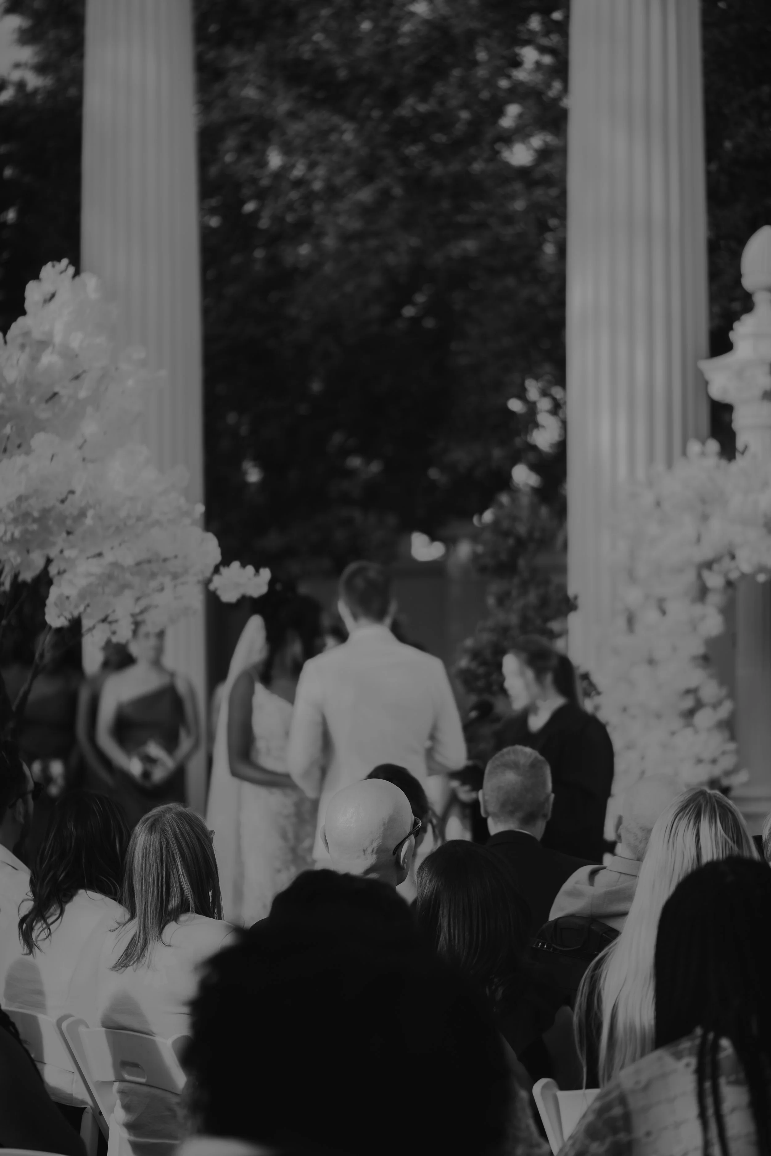 People gathered outdoors, seated in white chairs, facing a stage with two large columns and flowers on either side, with a man and woman standing on the stage.