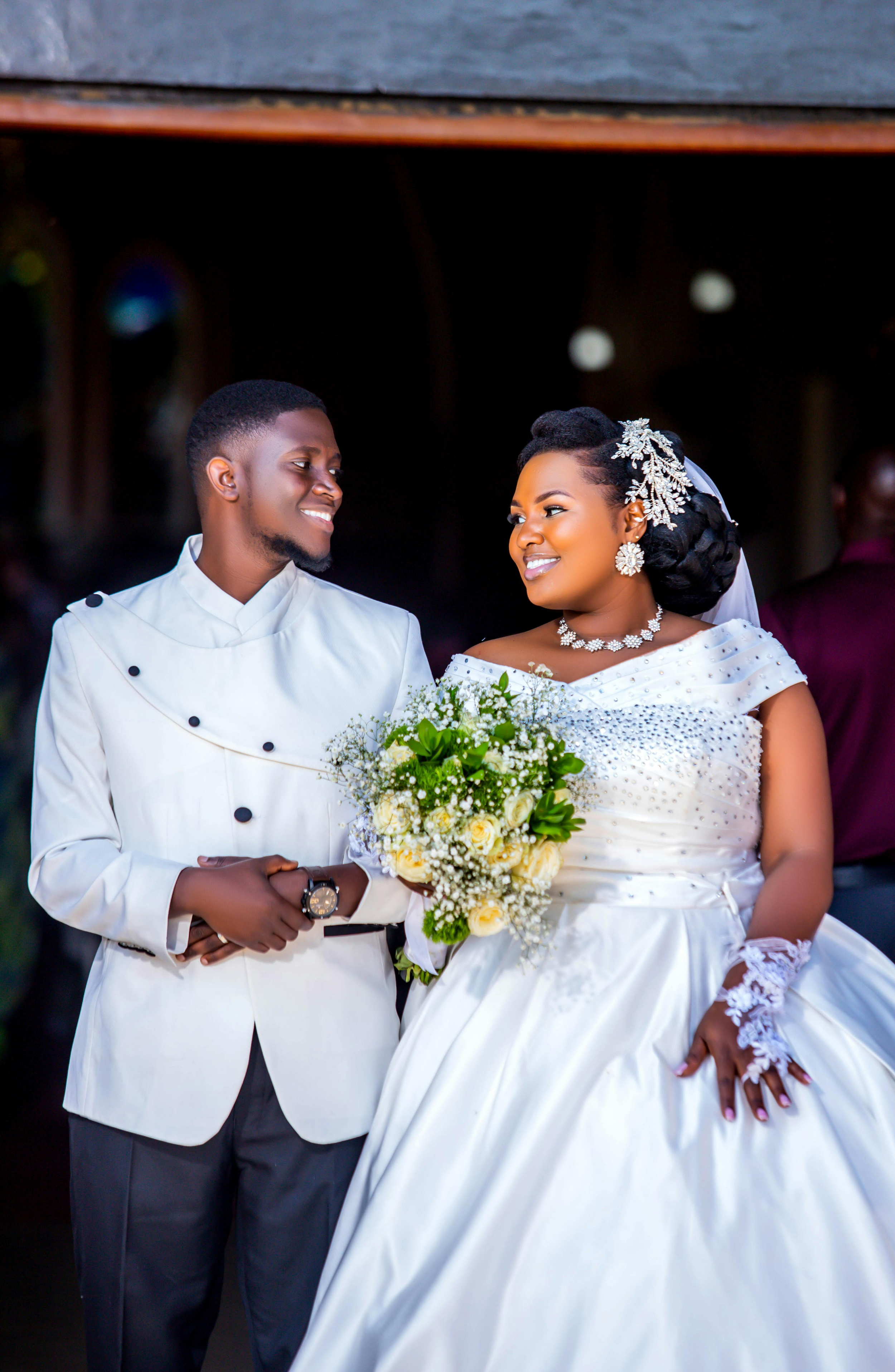 A man and woman dressed in wedding attire, holding hands and smiling at each other, with the woman carrying a bouquet of flowers.
