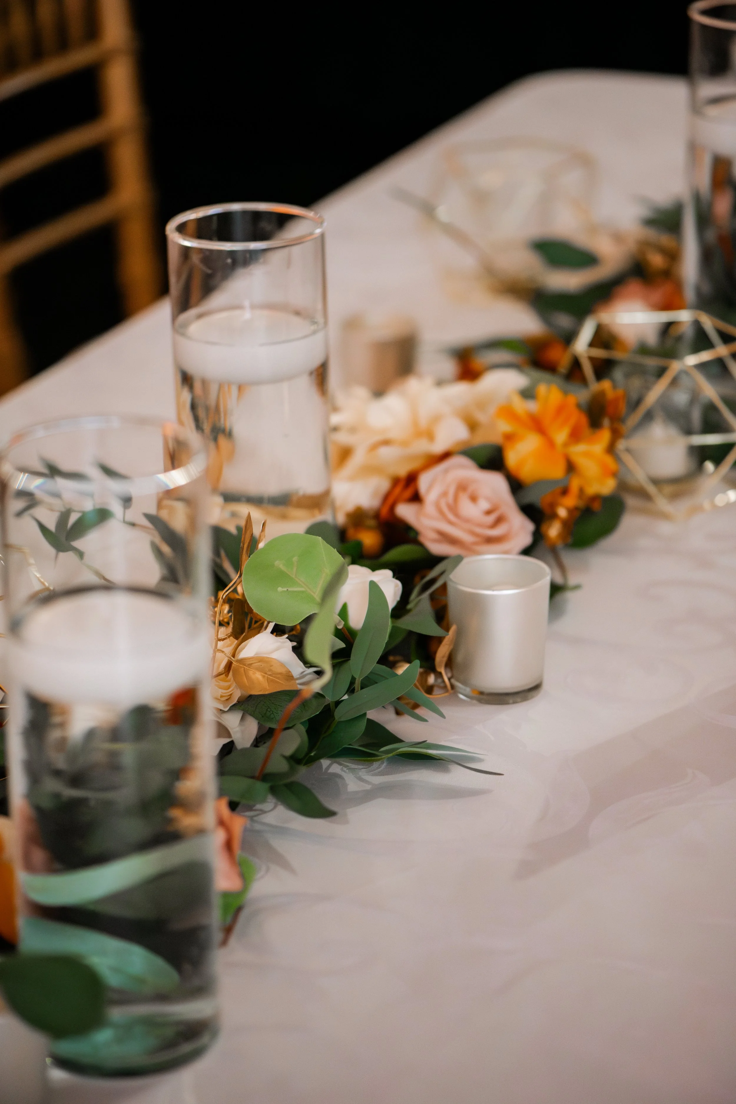 A table decorated with glass candle holders, a glass of water, and a floral centerpiece with peach and white flowers, green leaves, and decorative gold accents.