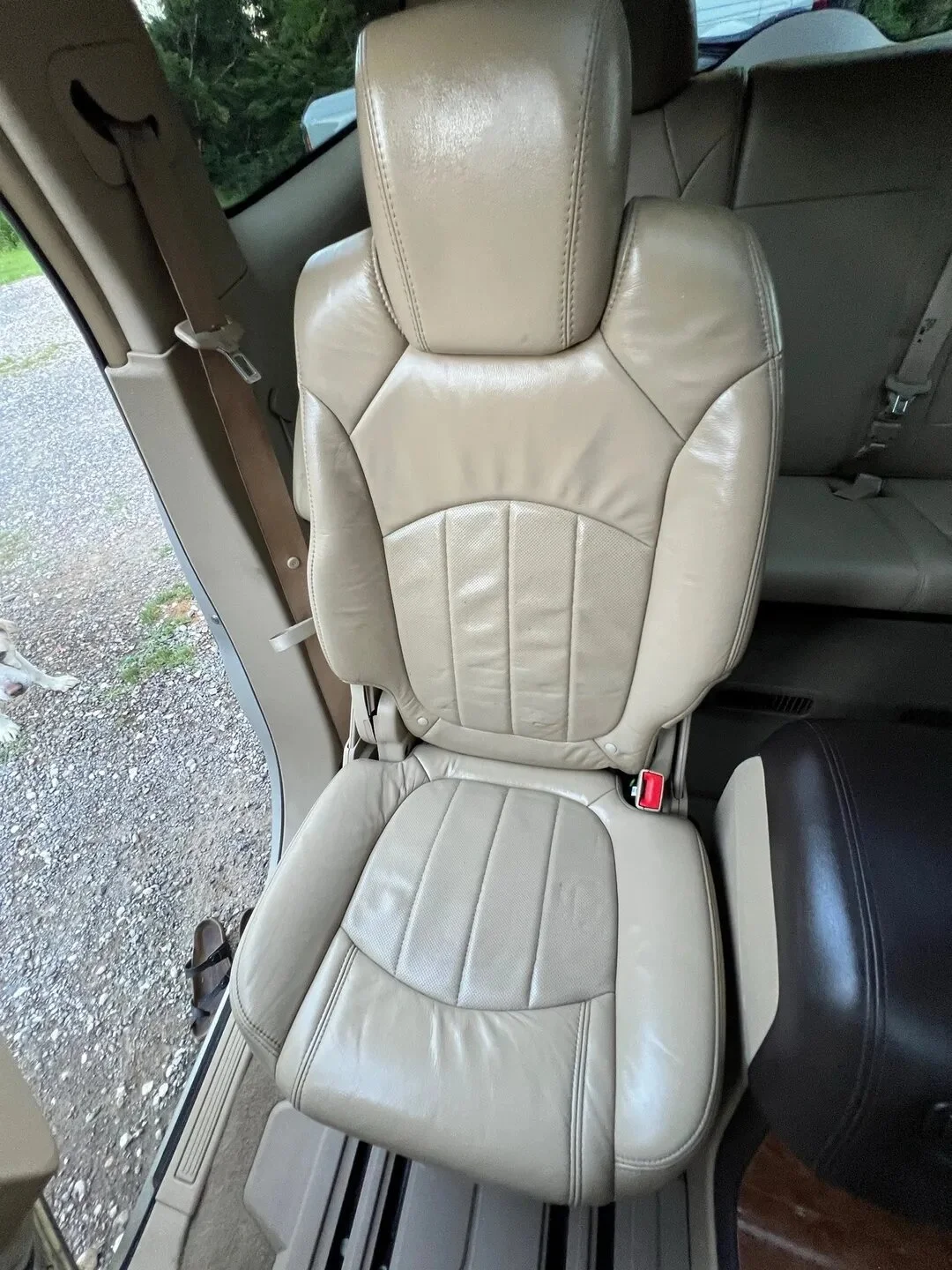 A beige leather vehicle seat in a car, with a headrest and seatbelt, next to a window showing gravel ground outside.