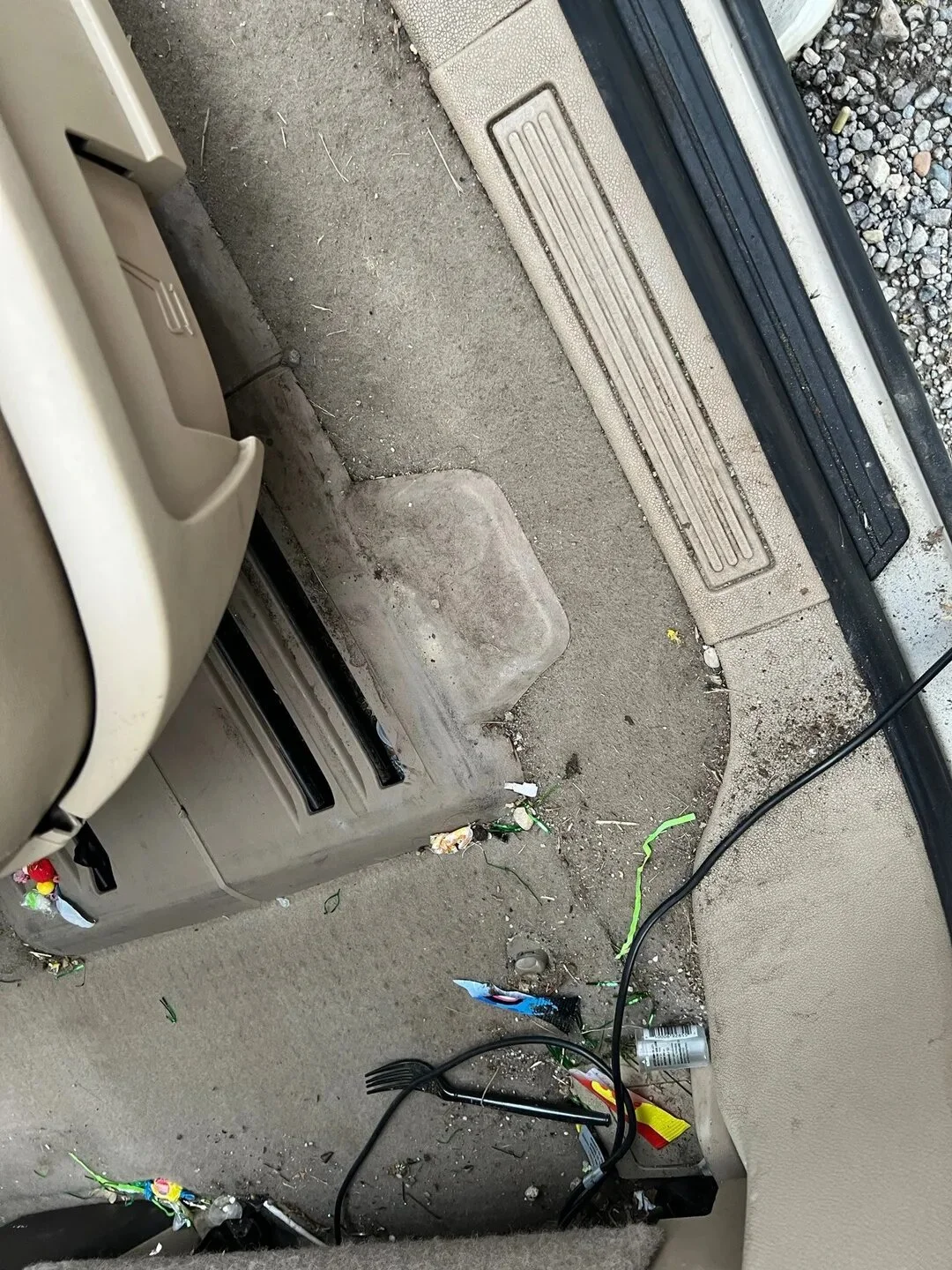 The interior of a car showing the footwell area with dirt, trash, cigarette butts, candy wrappers, a fork, a can, a cord, and a dirty floor mat.