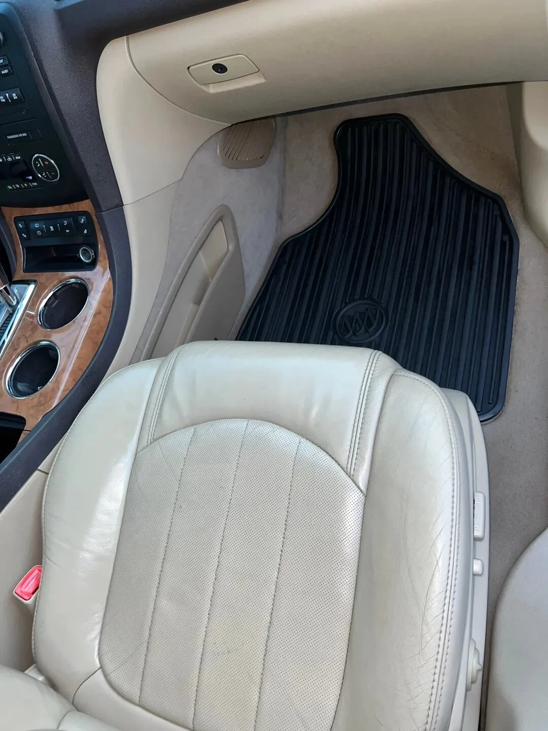 The interior of a car rear passenger area showing a beige leather seat, black rubber floor mat, part of the dashboard, and beige car floor with wood trim and controls.