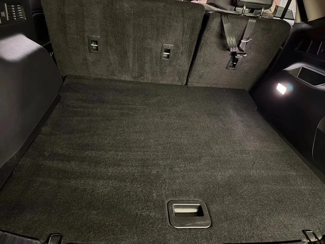 The image shows the interior of a vehicle's trunk or cargo area with a flat, clean, dark carpeted surface, and the back seats are upright.