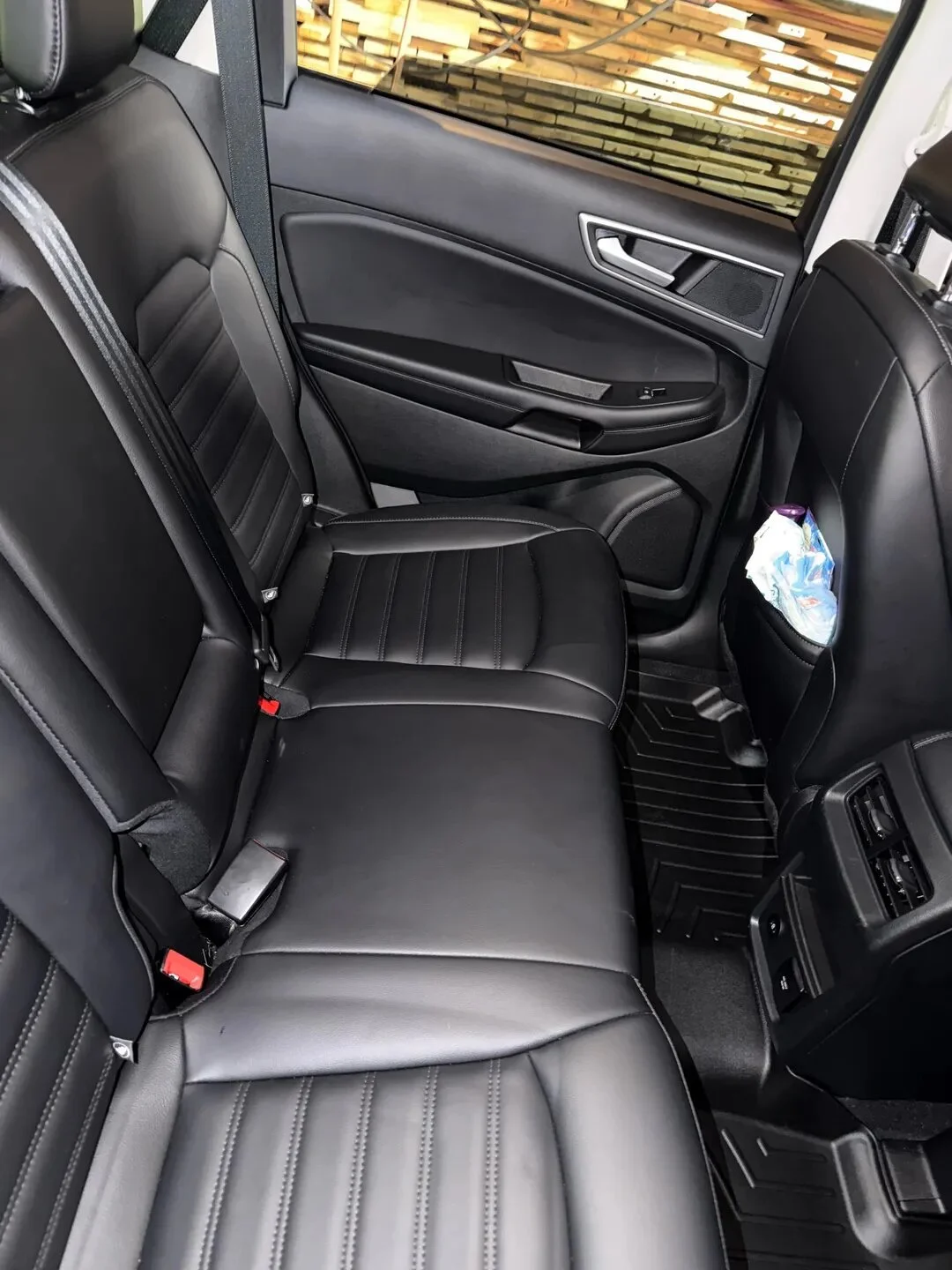 The back seat of a car with black leather seats, a side window with a wooden fence outside, and a back pocket with a pack of tissues.