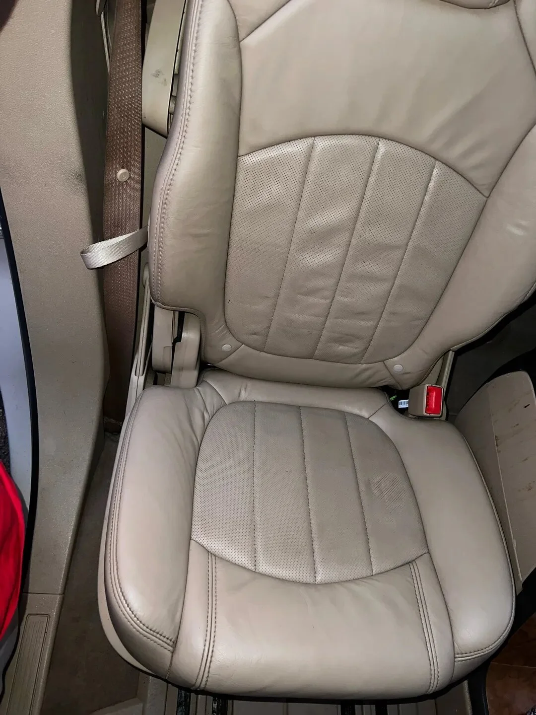 A beige leather airplane seat with vertical stitching, seen from above, featuring a seatbelt on the left and a red seatbelt buckle on the right.