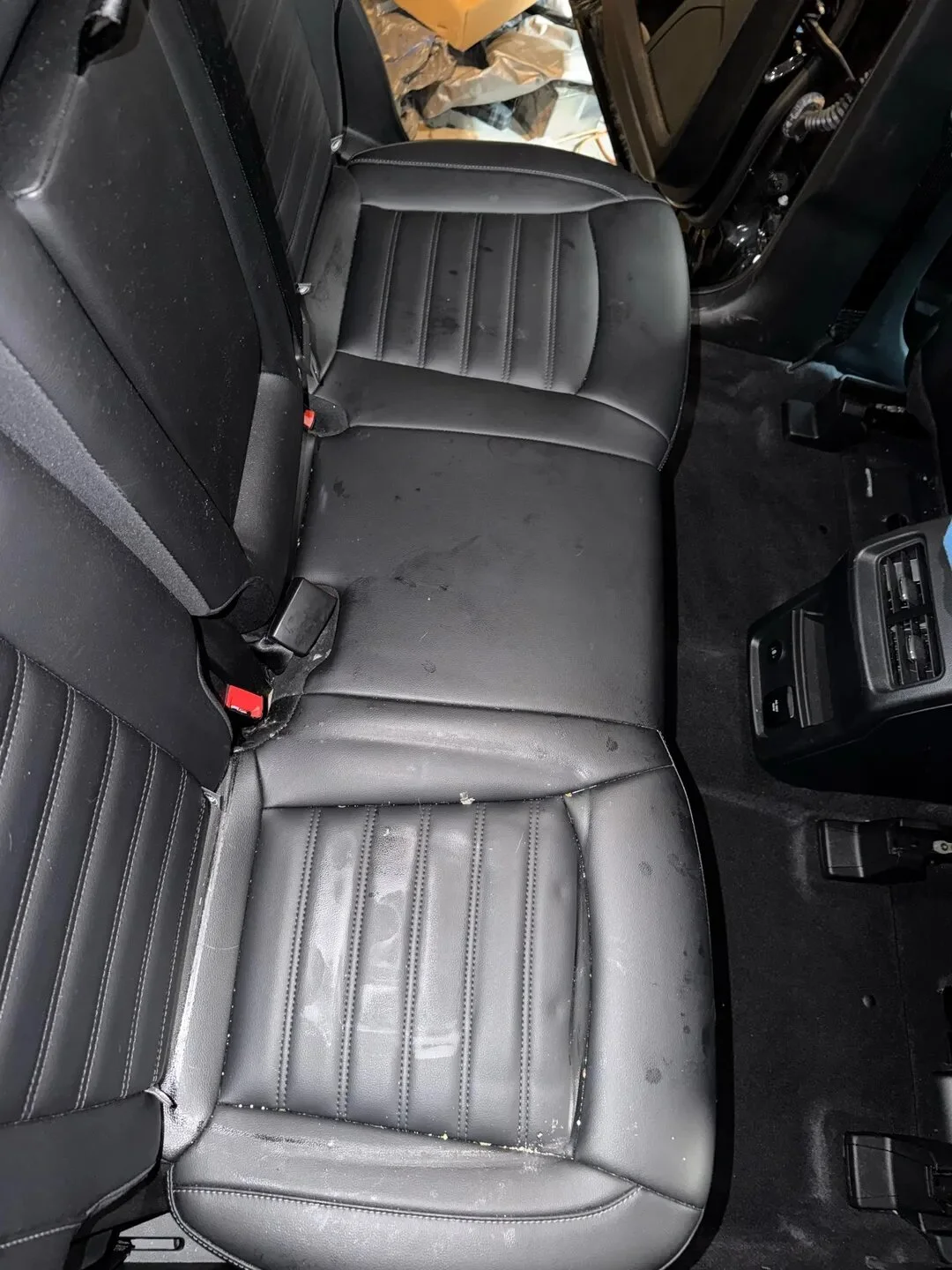 The back seat of a vehicle with black leather upholstery, showing signs of dirt and small stains, with part of the front dashboard and open door visible.
