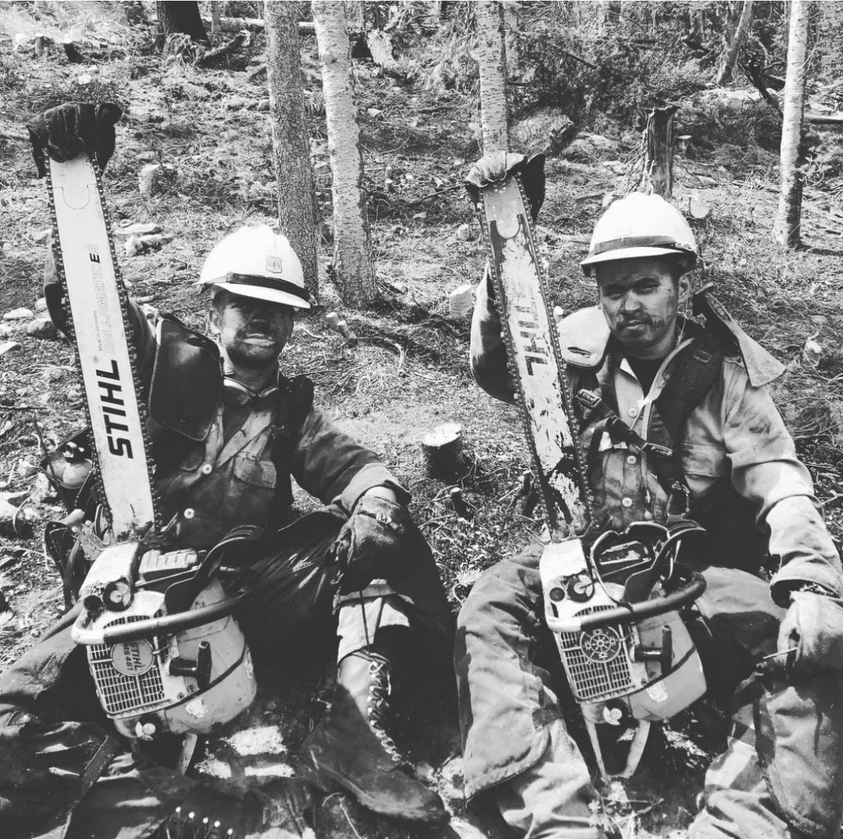 Two miners sitting in a forest, holding chainsaw blades, wearing helmets and protective gear.