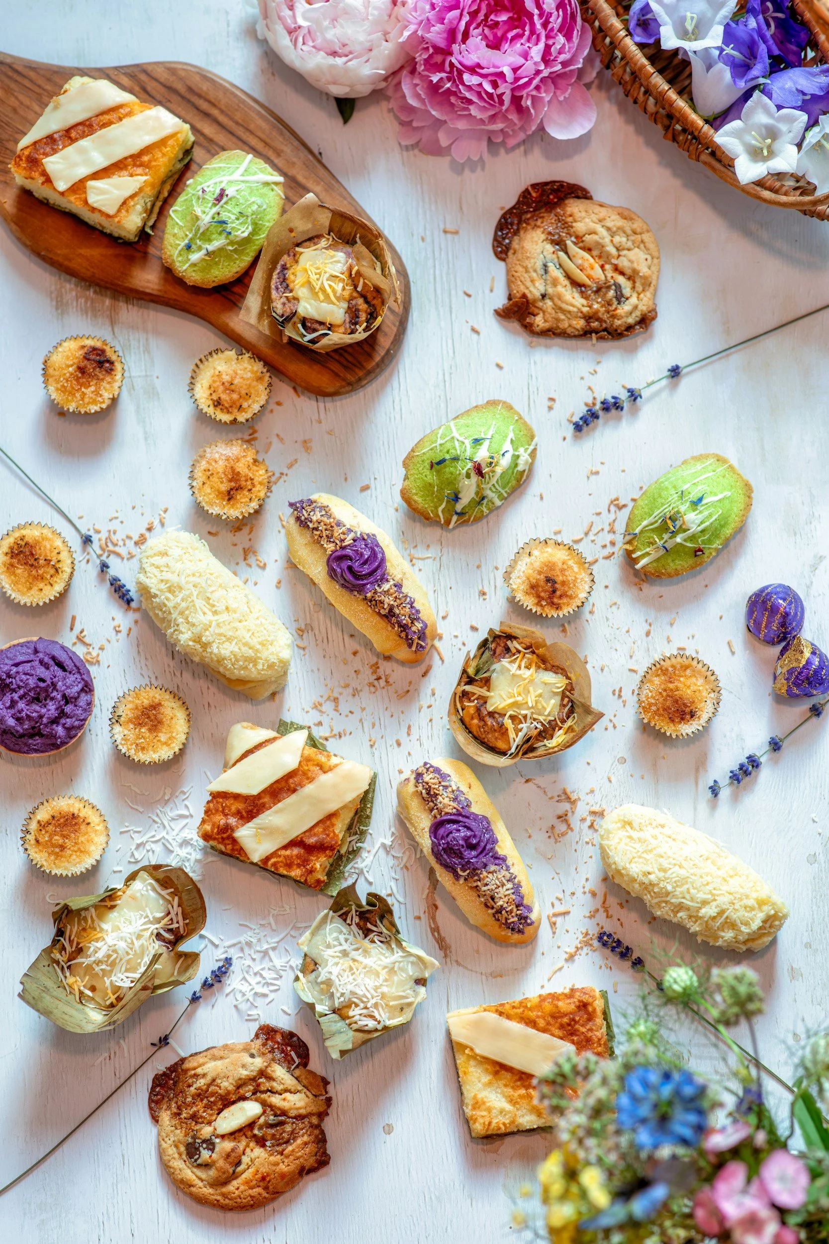 Assorted baked goods including cookies, cupcakes, and slices of cake placed on a white wooden surface, with pink, purple, and white flowers in the background.