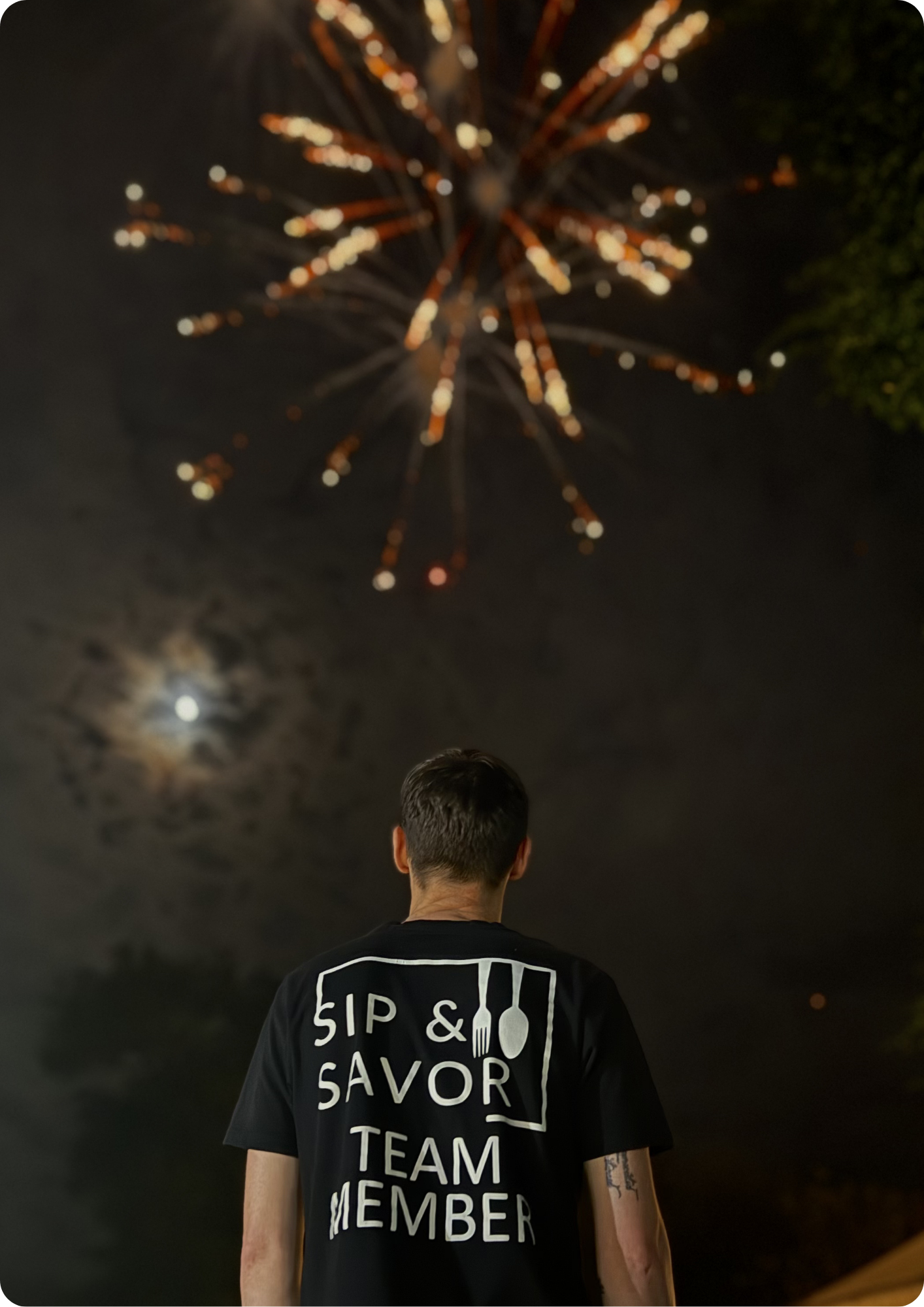 A person with short dark hair wearing a black T-shirt with the words 'Sip & Savor' and 'Team Member' printed on the back, standing in a dark environment looking up at fireworks lighting up the night sky.