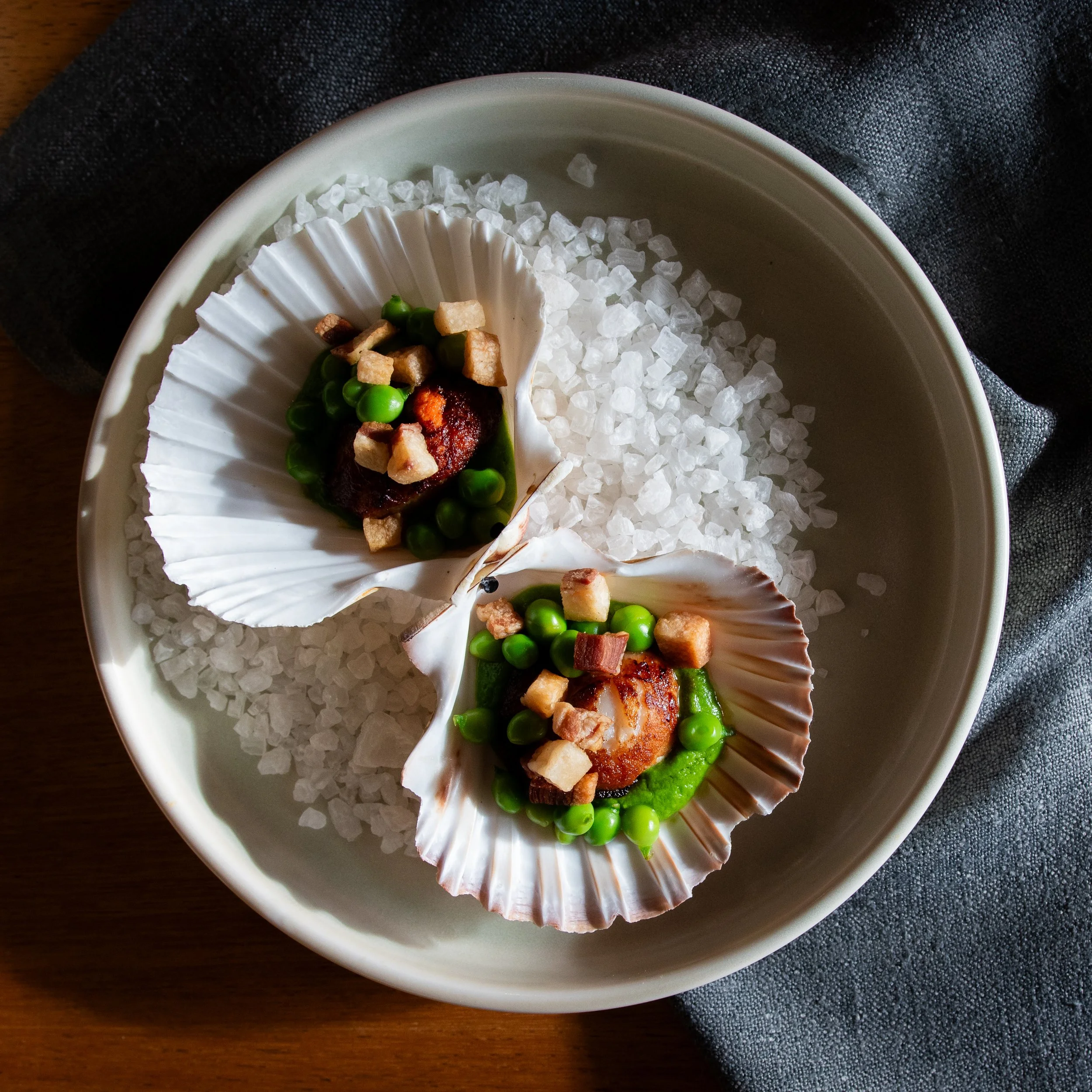 A ceramic bowl filled with coarse sea salt, with a scallop shell lined with green peas, croutons, and seared meat on top, resting on a dark textured cloth and a wooden surface.