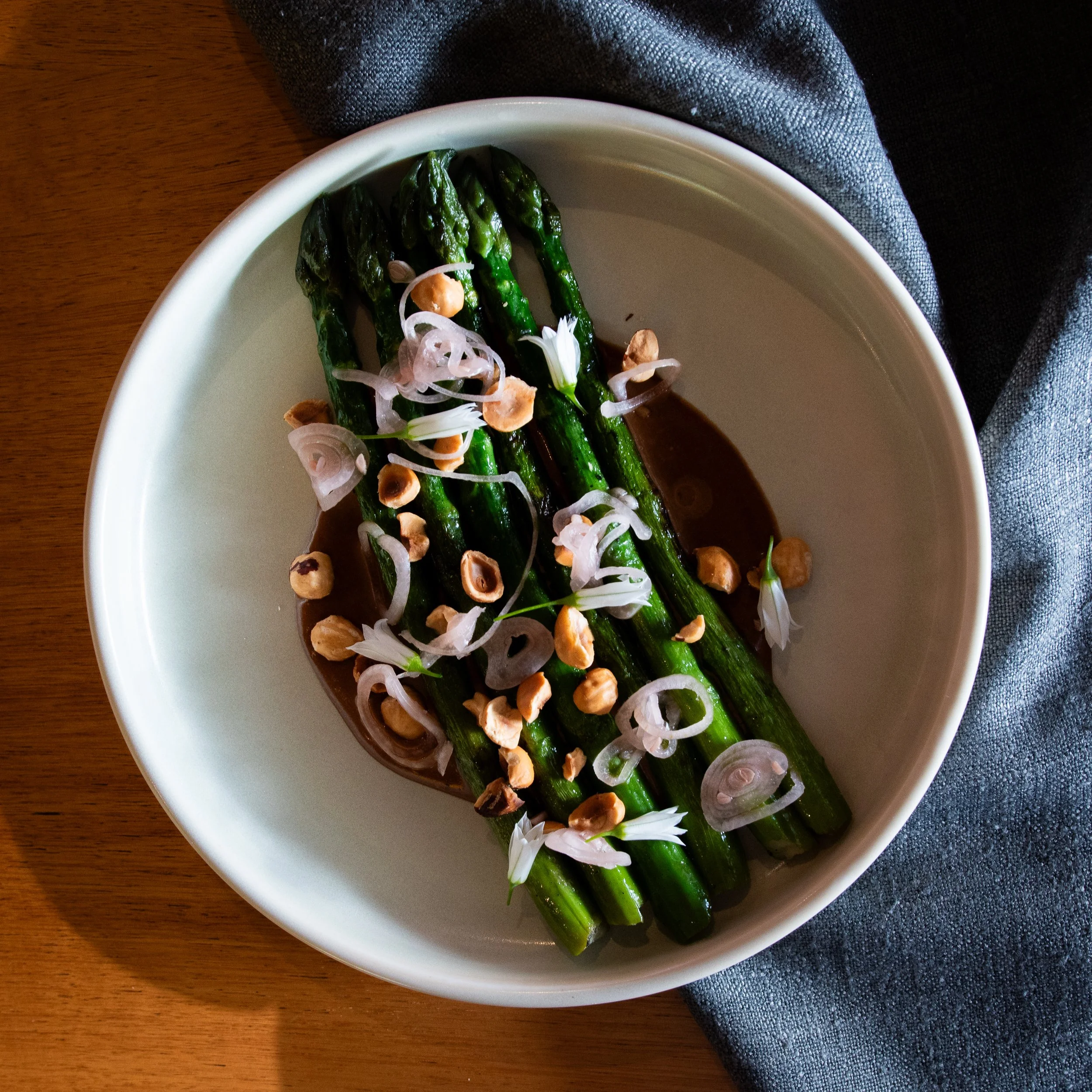 A white bowl containing grilled asparagus topped with sliced onions, chopped nuts, and drizzled with a dark sauce, placed on a wooden surface with a gray napkin underneath.
